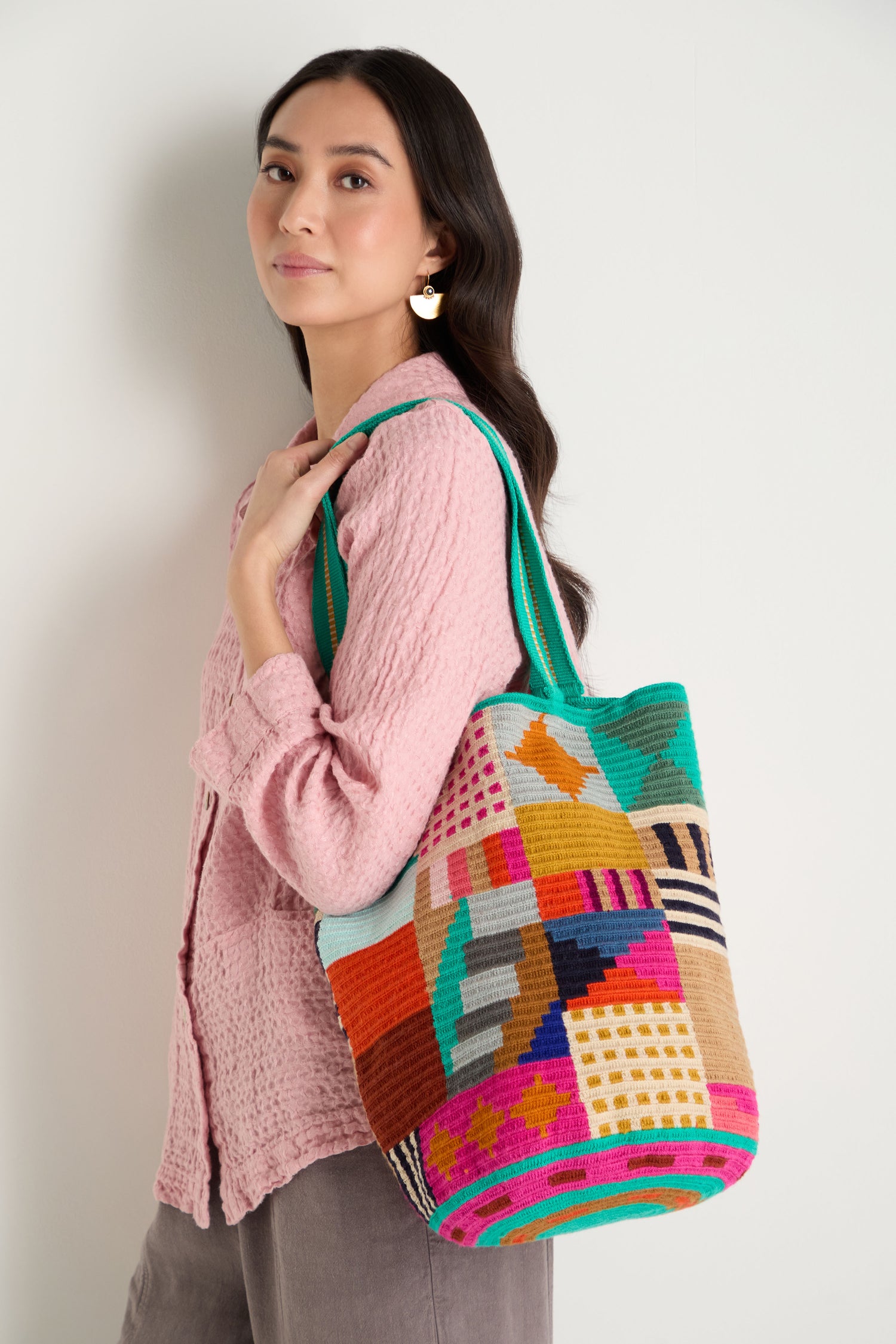 A woman stands against a plain light background, wearing a textured pink blouse and gray pants, carrying the Hand Crocheted Cala Tote Bag with a colorful geometric design over her shoulder.