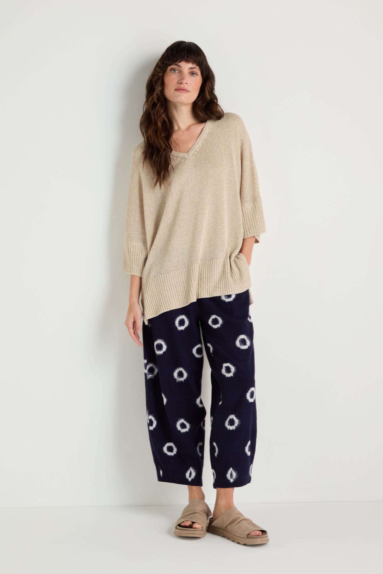 A woman stands against a white wall wearing the V Neck Relaxed Knit Putty Top, navy pants with white circular patterns, and beige sandals.