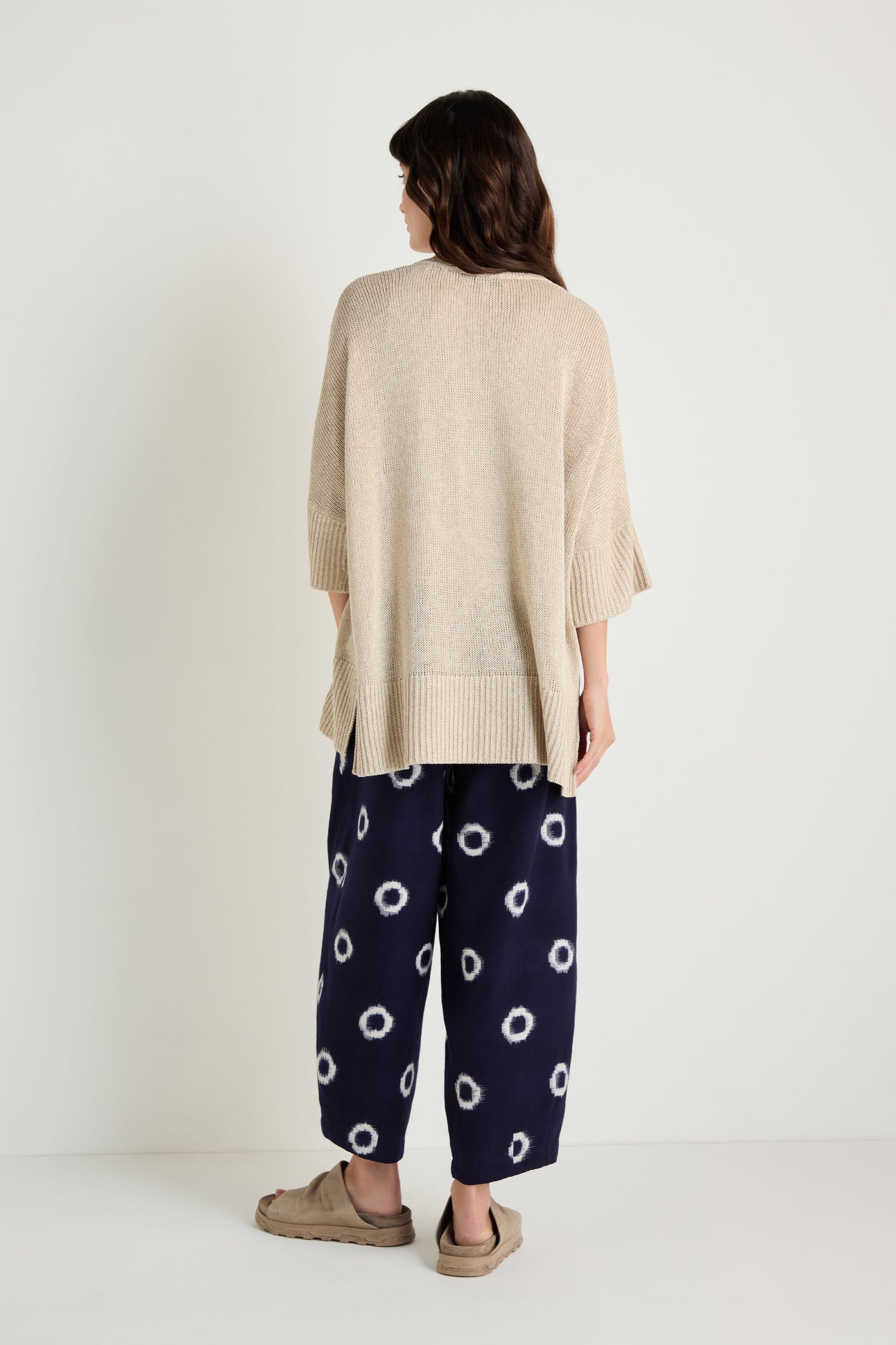 A person stands facing away, wearing a V Neck Relaxed Knit Putty Top, navy blue pants with white circular patterns, and tan slip-on shoes against a plain white background.