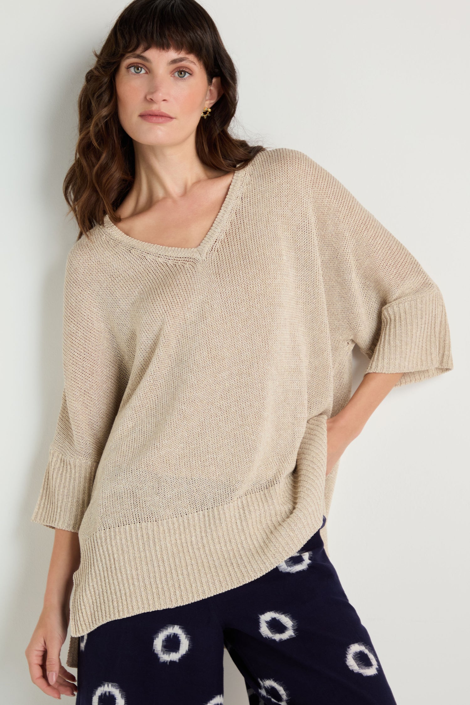 A woman wearing a V Neck Relaxed Knit Putty Top with elbow-length sleeves and navy pants featuring a white circular pattern stands against a plain background.