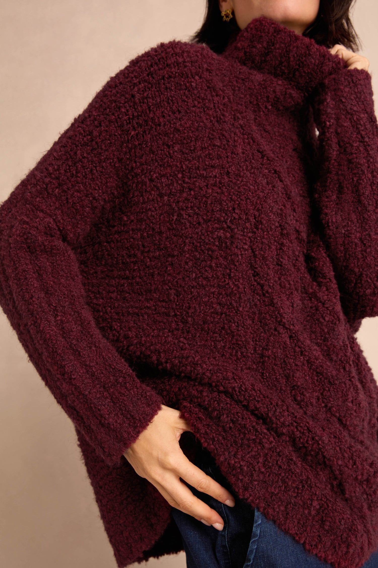 Wearing the Bordeaux Boucle Cable Knit Jumper—a textured, dark maroon piece crafted in Italy—paired with blue jeans, one hand on the hip against a neutral background; a perfect example of elegant winter knitwear.