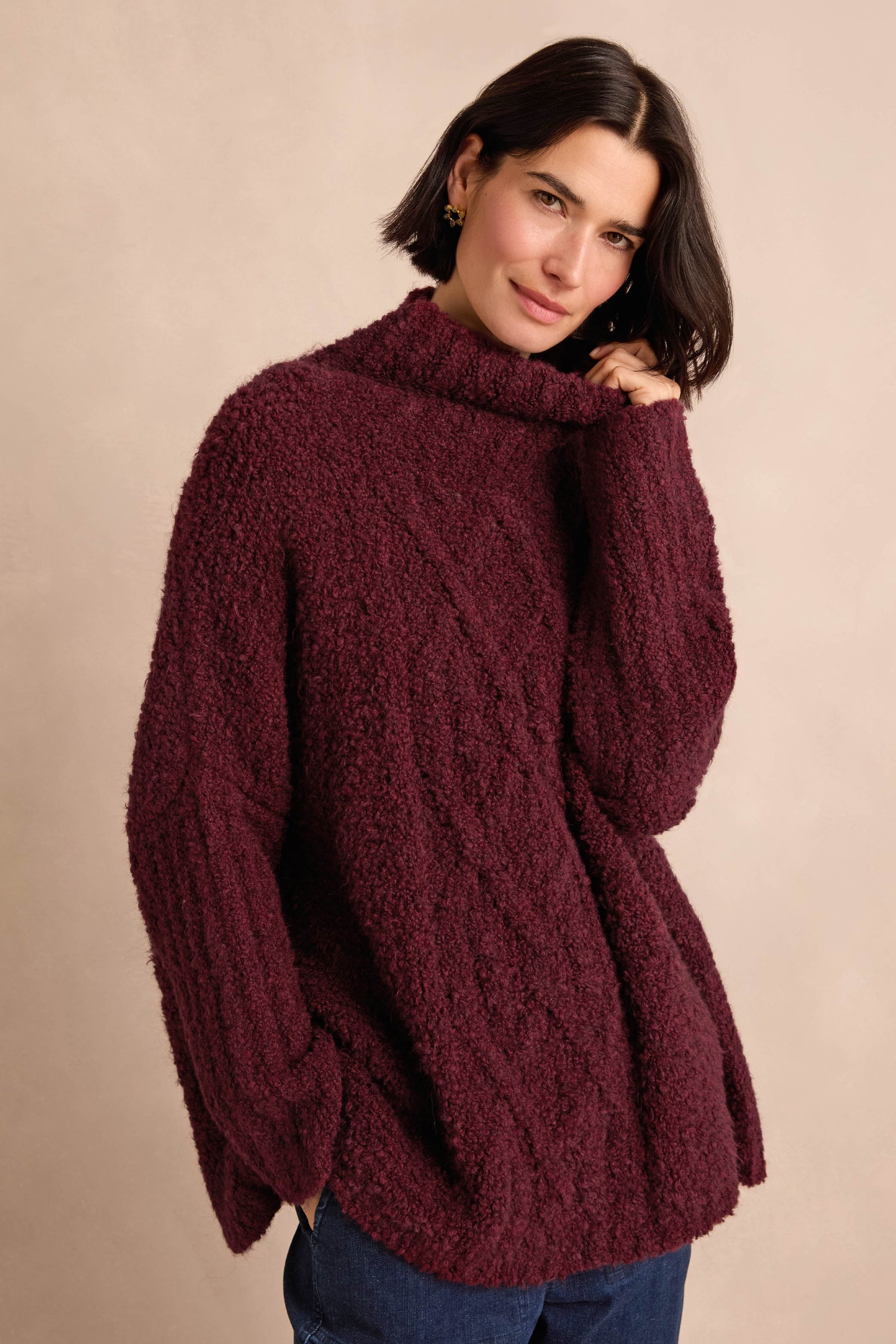Wearing the Bordeaux Boucle Cable Knit Jumper—an oversized, textured turtleneck knit in a rich burgundy hue—this individual stands against a beige background, gently touching the collar and sporting a relaxed expression. Made in Italy, this cozy winter staple offers both style and comfort.