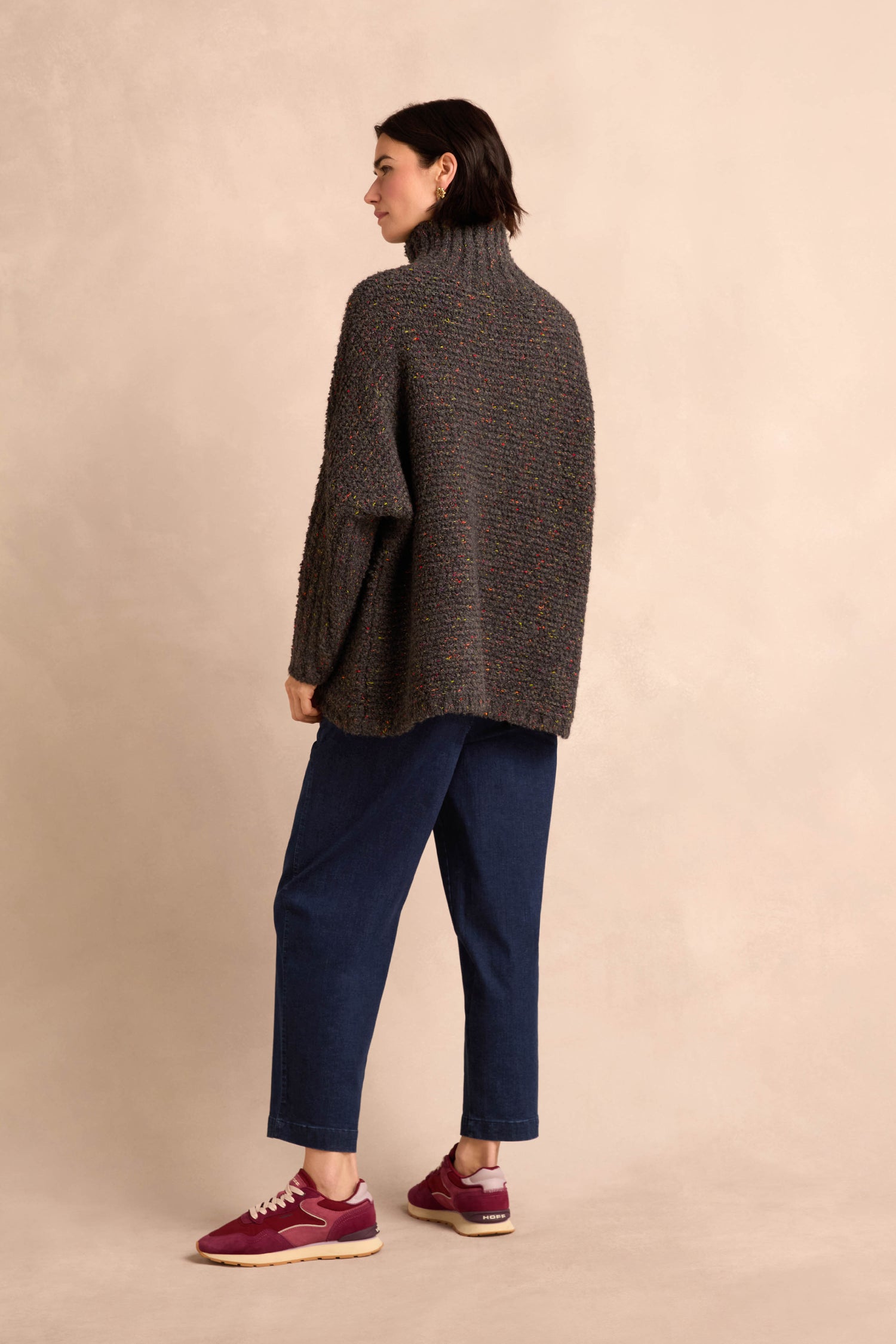 A person stands sideways against a plain beige background wearing the Graphite Boucle Cable Knit Jumper—a chunky gray turtleneck sweater with a soft textured finish—paired with loose blue jeans and maroon sneakers.