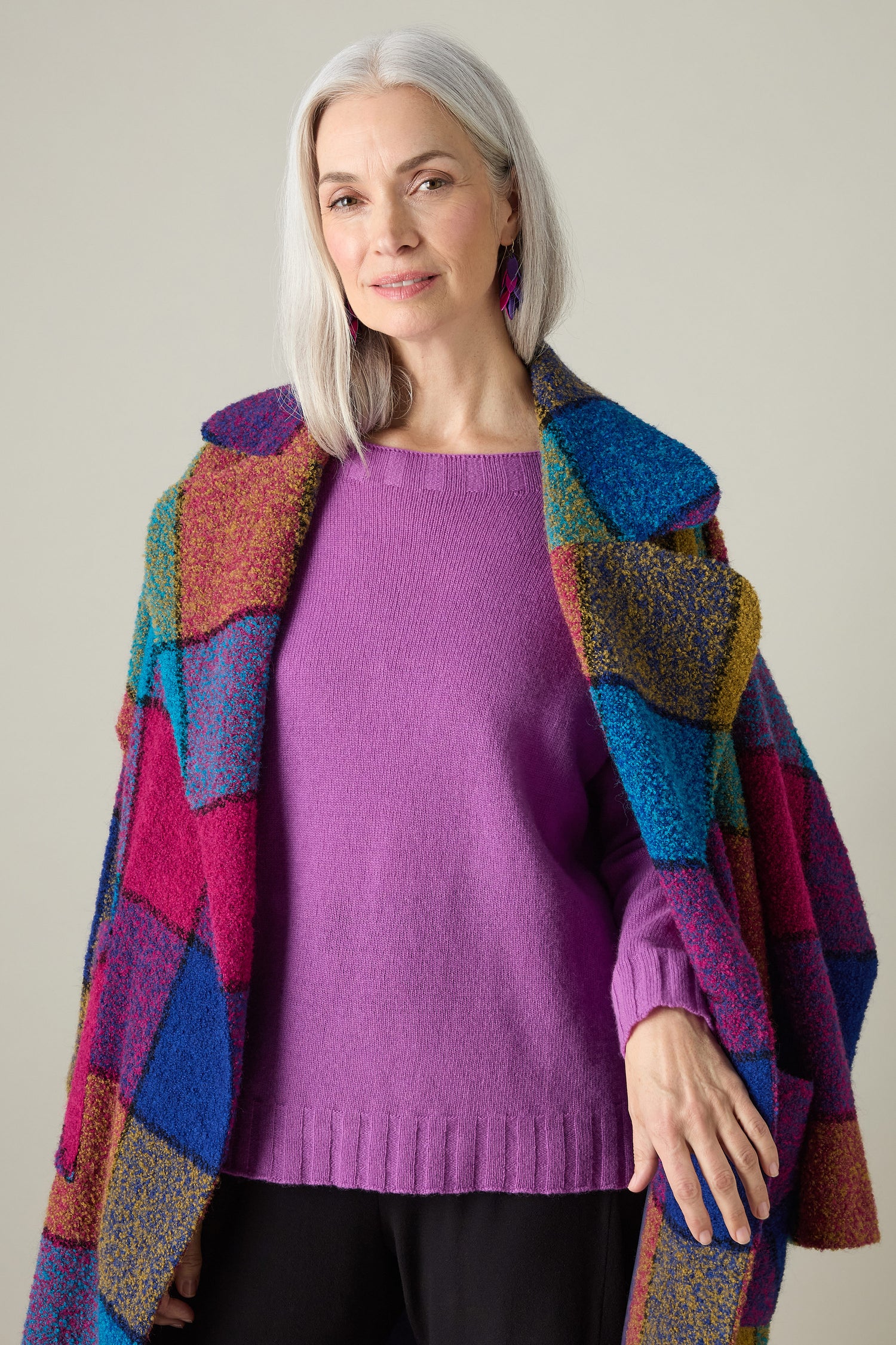 A woman with gray hair wears a colorful patchwork coat and a purple Merino Wool Crop Jumper, exuding luxurious comfort while standing against a plain background.