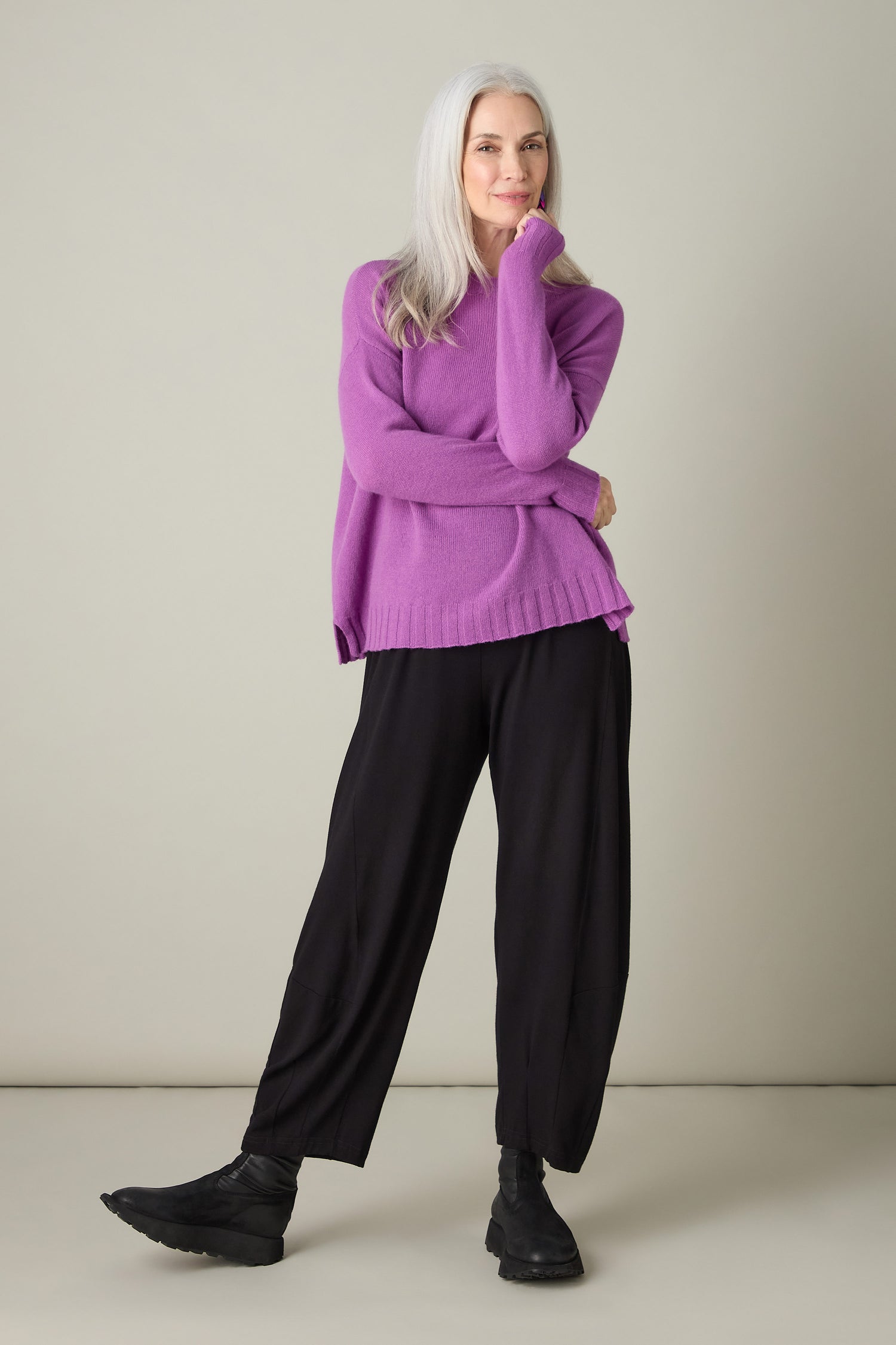 A person with long gray hair, wearing a purple Merino Wool Crop Jumper and black pants, stands against a plain background. Smiling with luxurious comfort, they place one hand on their chin.