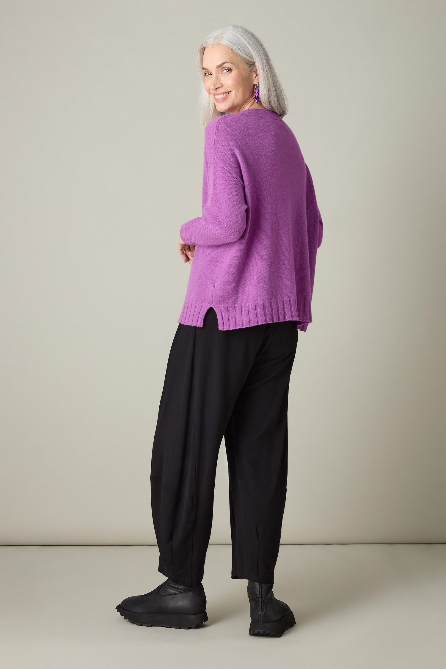 A woman with long white hair, dressed in a luxurious Merino Wool Crop Jumper in purple and black pants, faces slightly sideways as she looks back and smiles.
