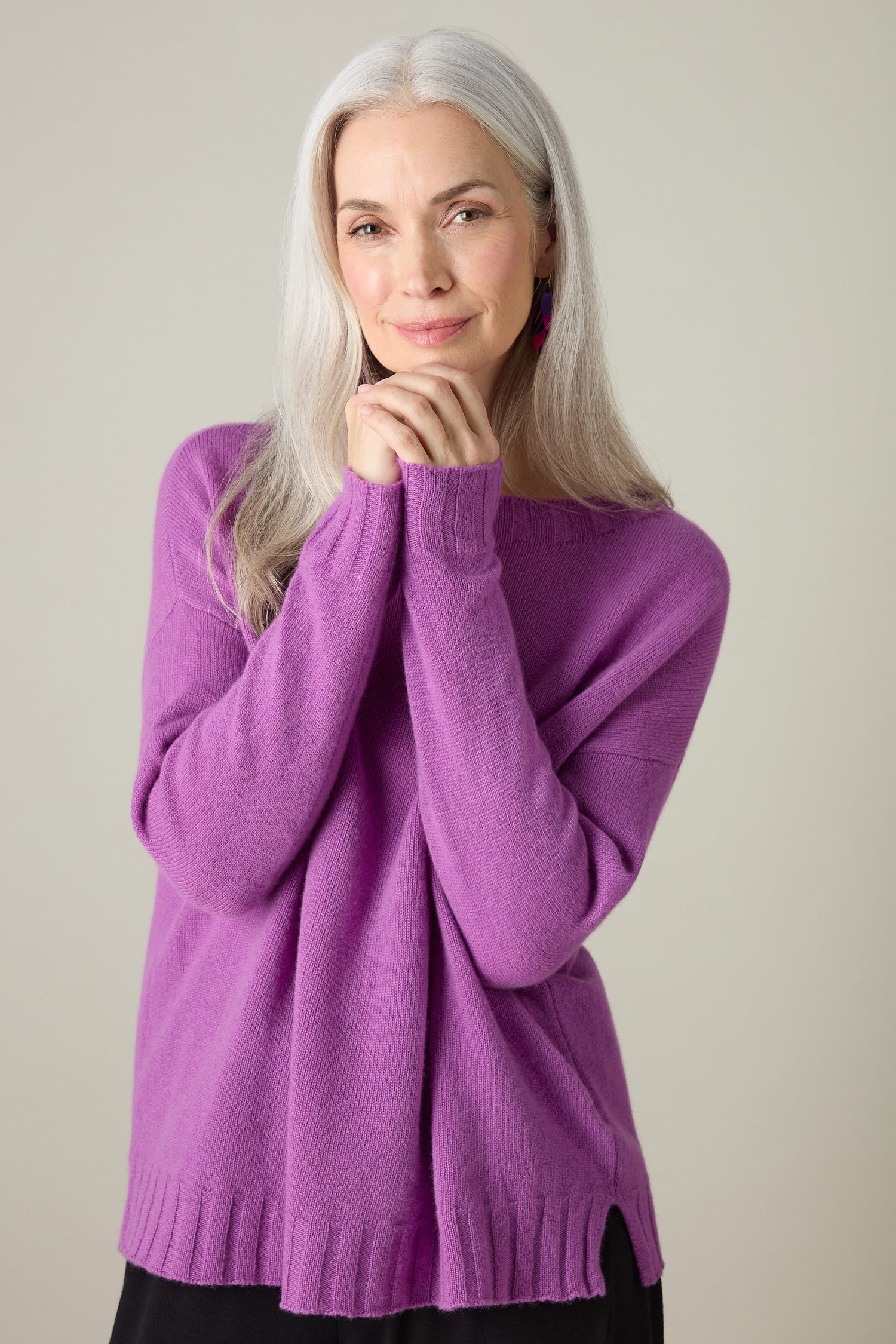 A person with long gray hair is wearing a luxurious Merino Wool Crop Jumper in a rich purple hue, posing with hands gently clasped under their chin against a neutral background.