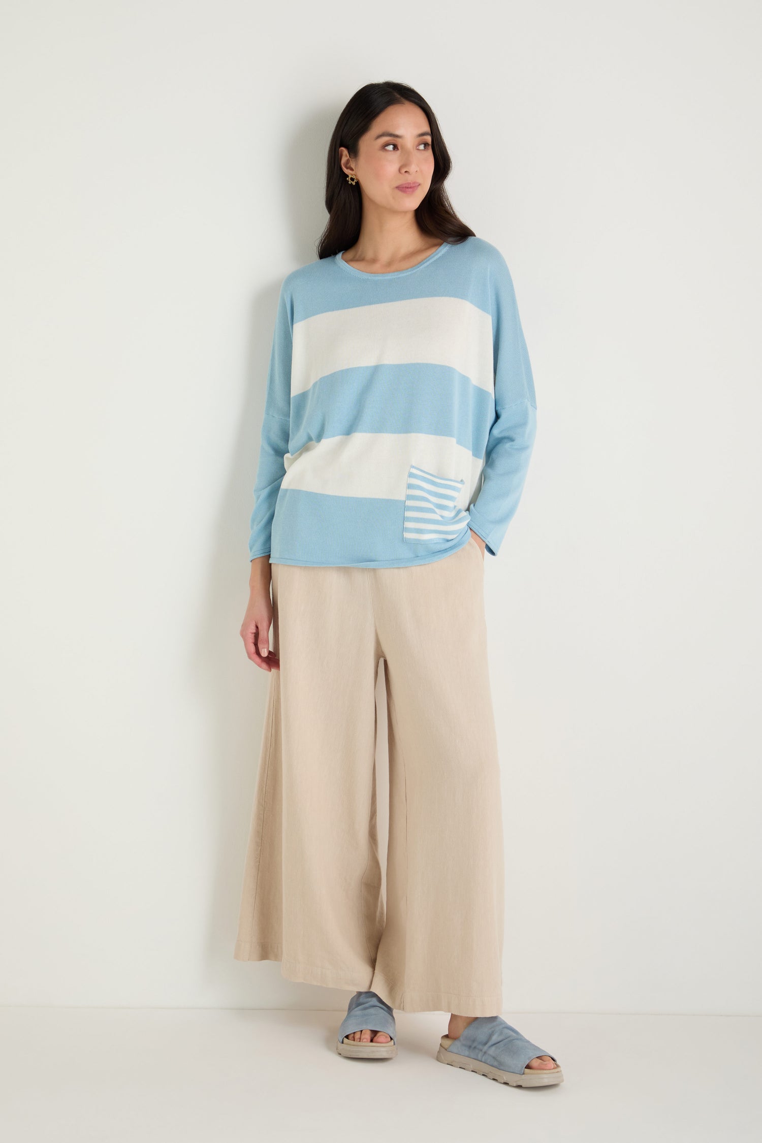 A woman stands against a white wall wearing the Stripe Pocket Knit Top in light blue and white stripes, paired with beige wide-leg pants and blue sandals.