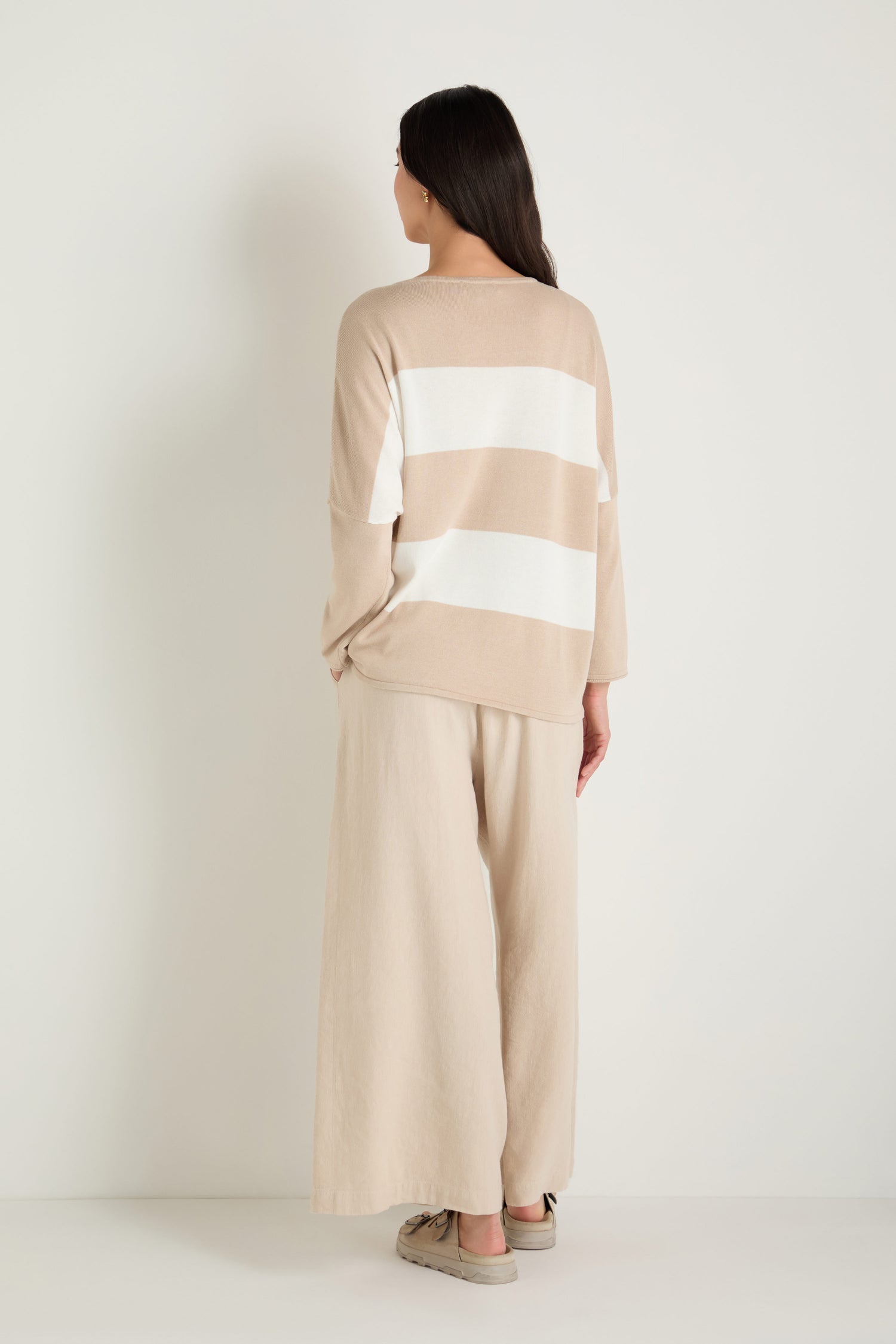 A woman stands against a white background, facing away. She wears a beige and white Stripe Pocket Knit Top with a relaxed boxy fit, wide-legged beige modal cotton pants, and beige shoes.