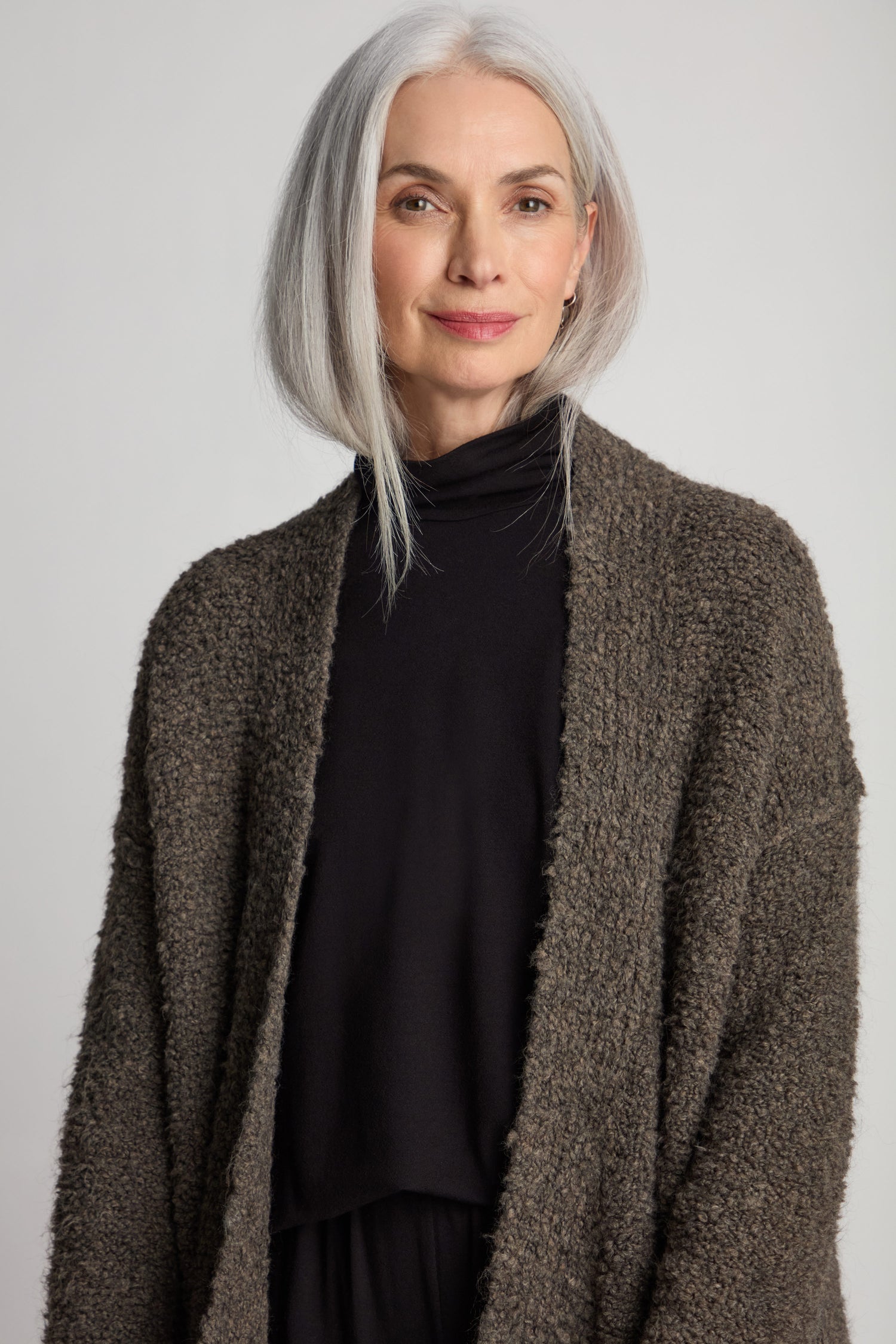 A woman with shoulder-length gray hair, wearing a black shirt and the Boucle Knit Long Cardi, stands facing the camera, smiling slightly.