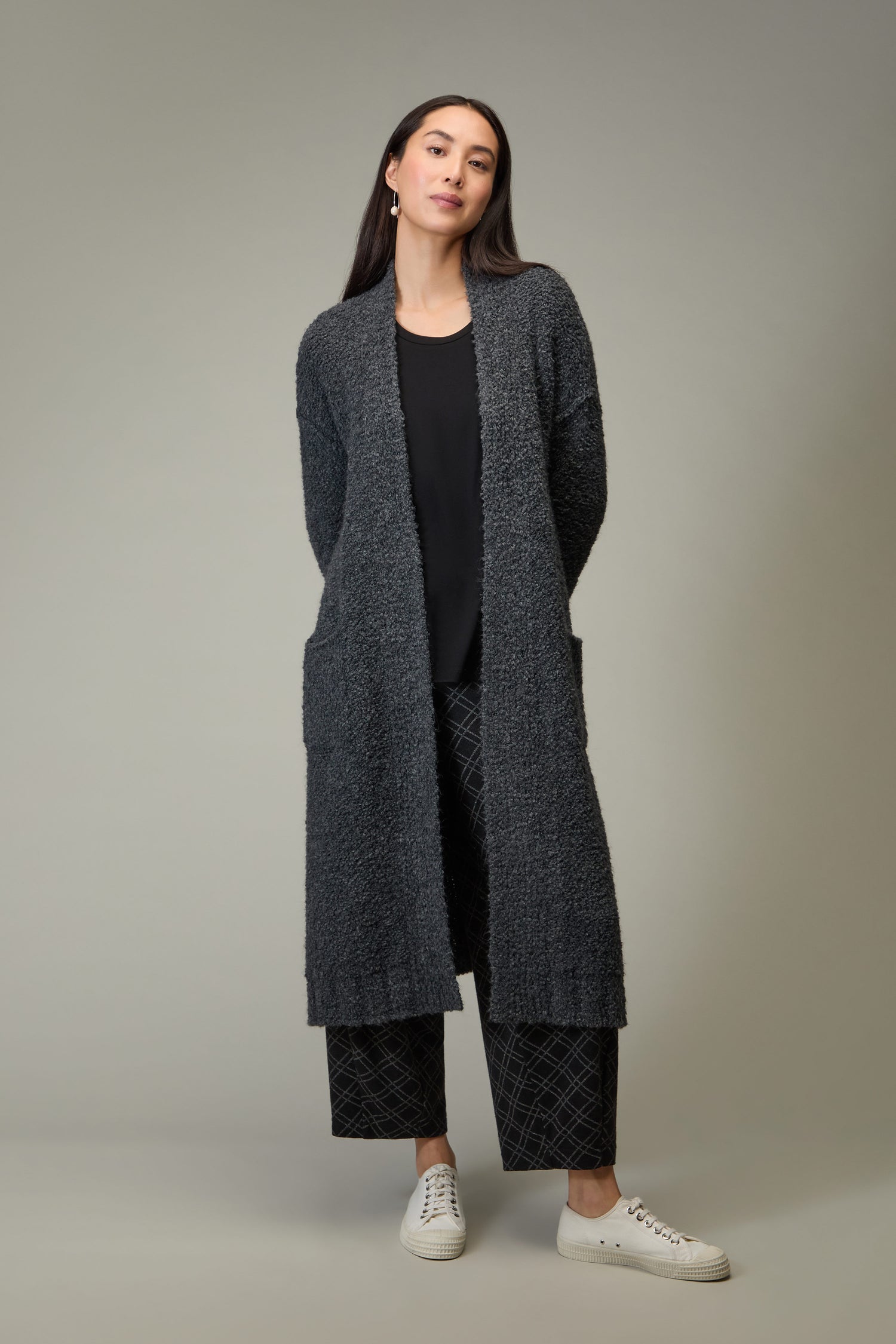 Woman stands against a plain background wearing the Charcoal Boucle Knit Long Cardi over a black top, patterned black pants, and white sneakers—an ideal look to elevate your autumn wardrobe.