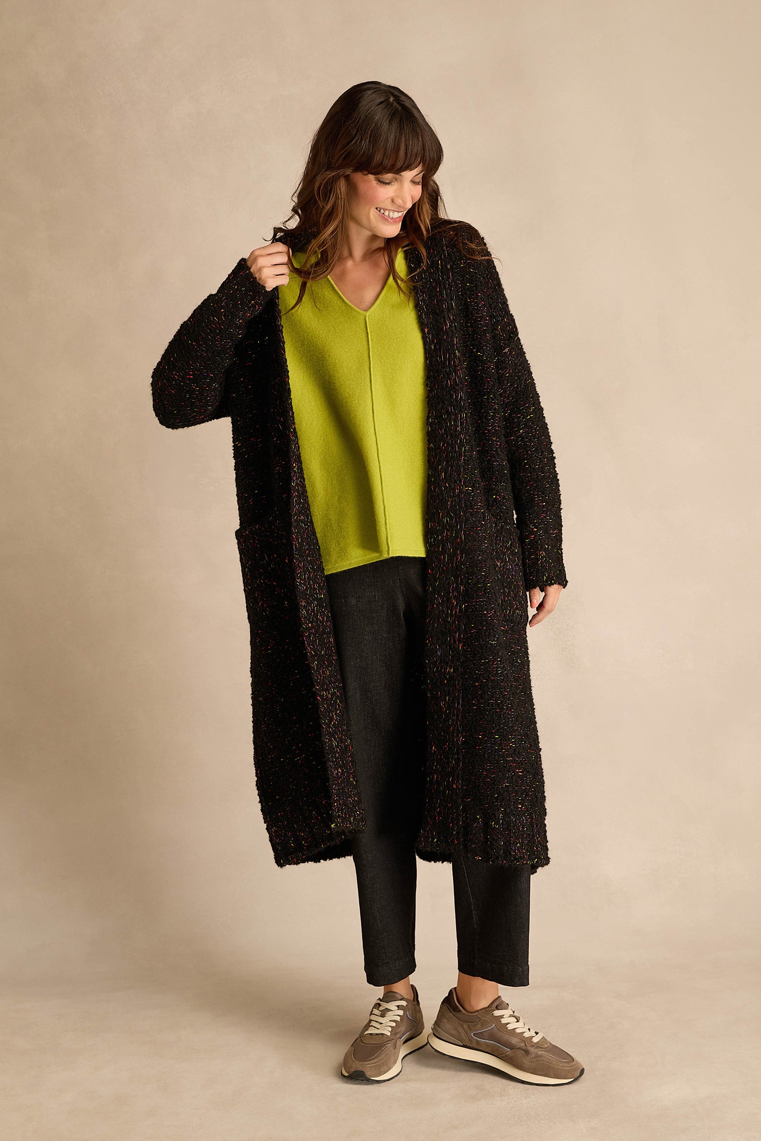 A woman stands smiling against a neutral background, wearing the Black Boucle Knit Long Line Speckle Cardi layered over a bright green V-neck top, paired with black pants and beige sneakers.