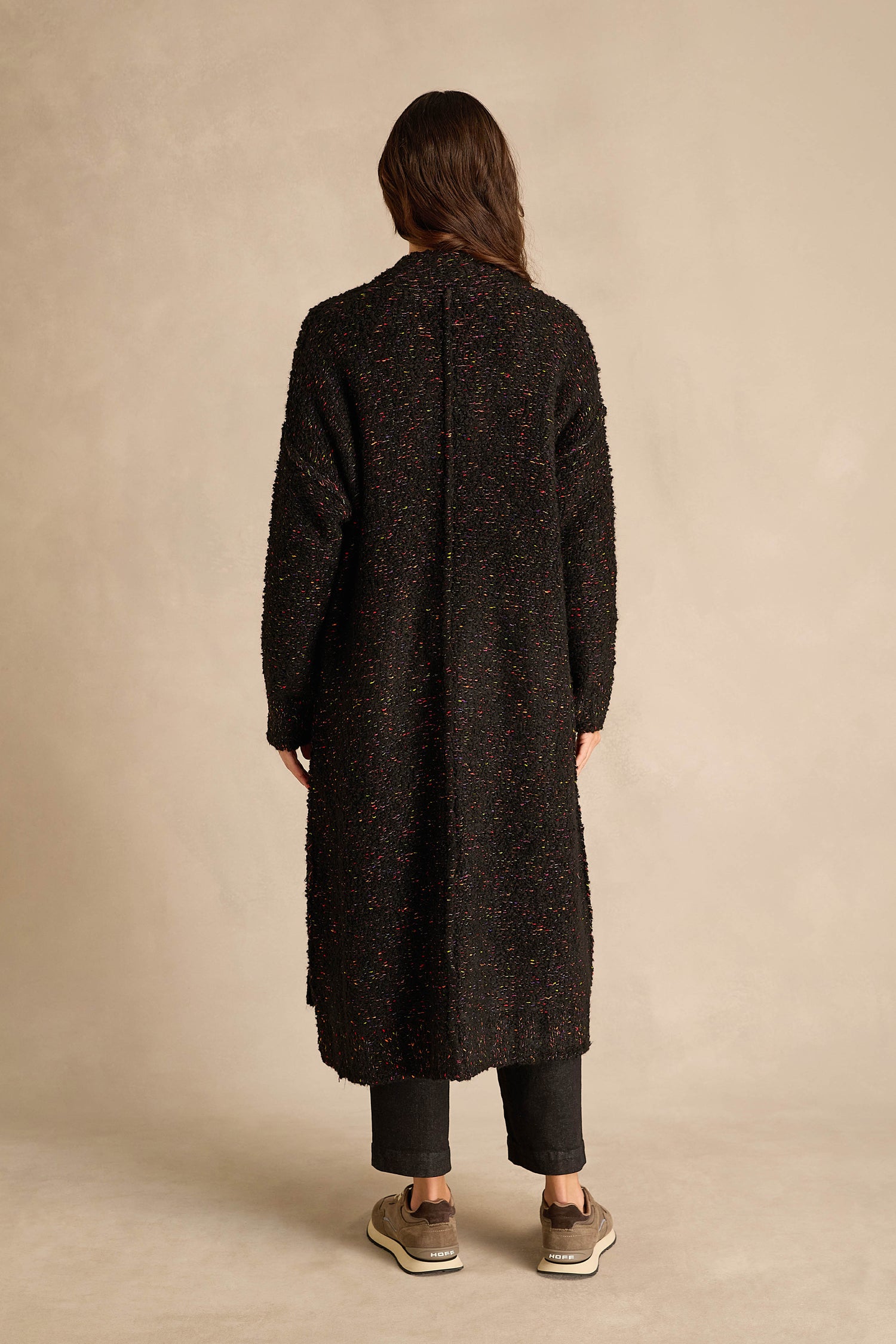 A person stands with their back to the camera, wearing the Black Boucle Knit Long Line Speckle Cardi, paired with dark pants and beige sneakers against a plain beige background.