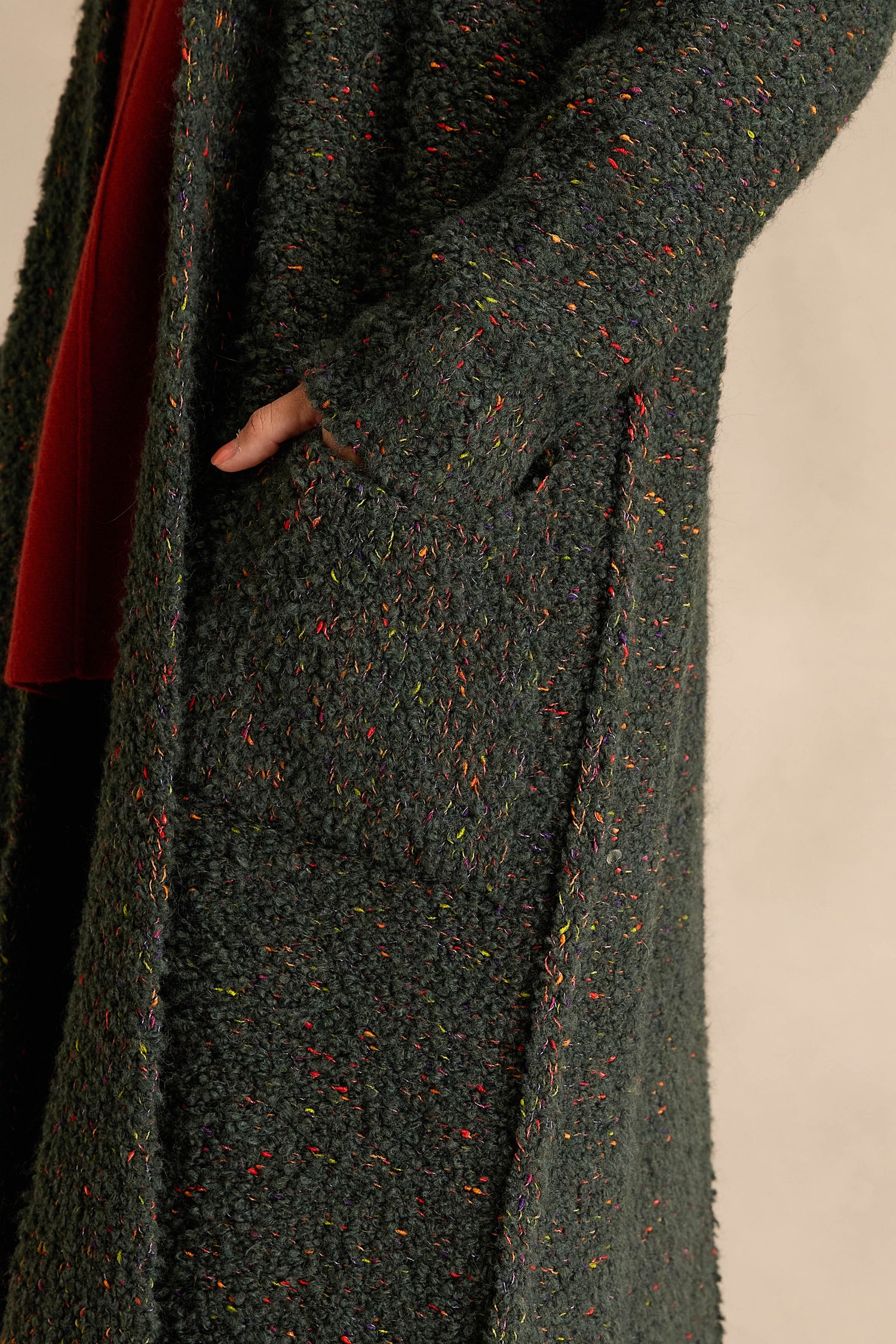 Someone wearing the Forest Boucle Knit Long Line Speckle Cardi, featuring a textured dark green knit with colorful flecks, has their hand tucked into one of its large front pockets.
