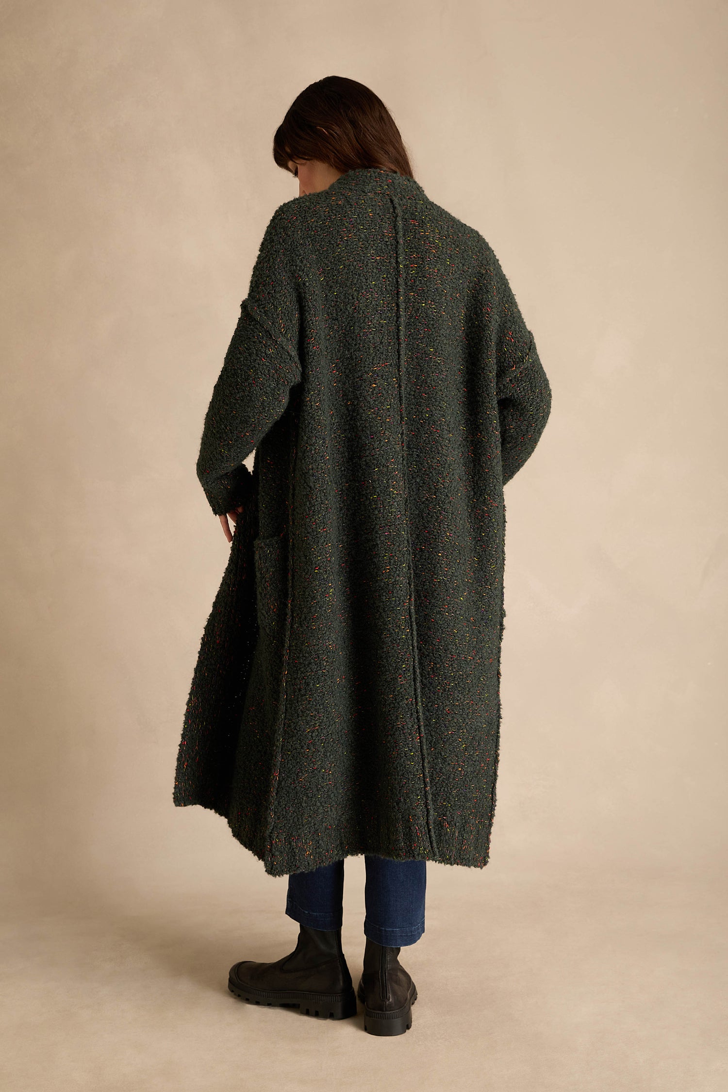 A person stands with their back to the camera, wearing the Forest Boucle Knit Long Line Speckle Cardi, paired with blue jeans and black boots, against a plain beige background.