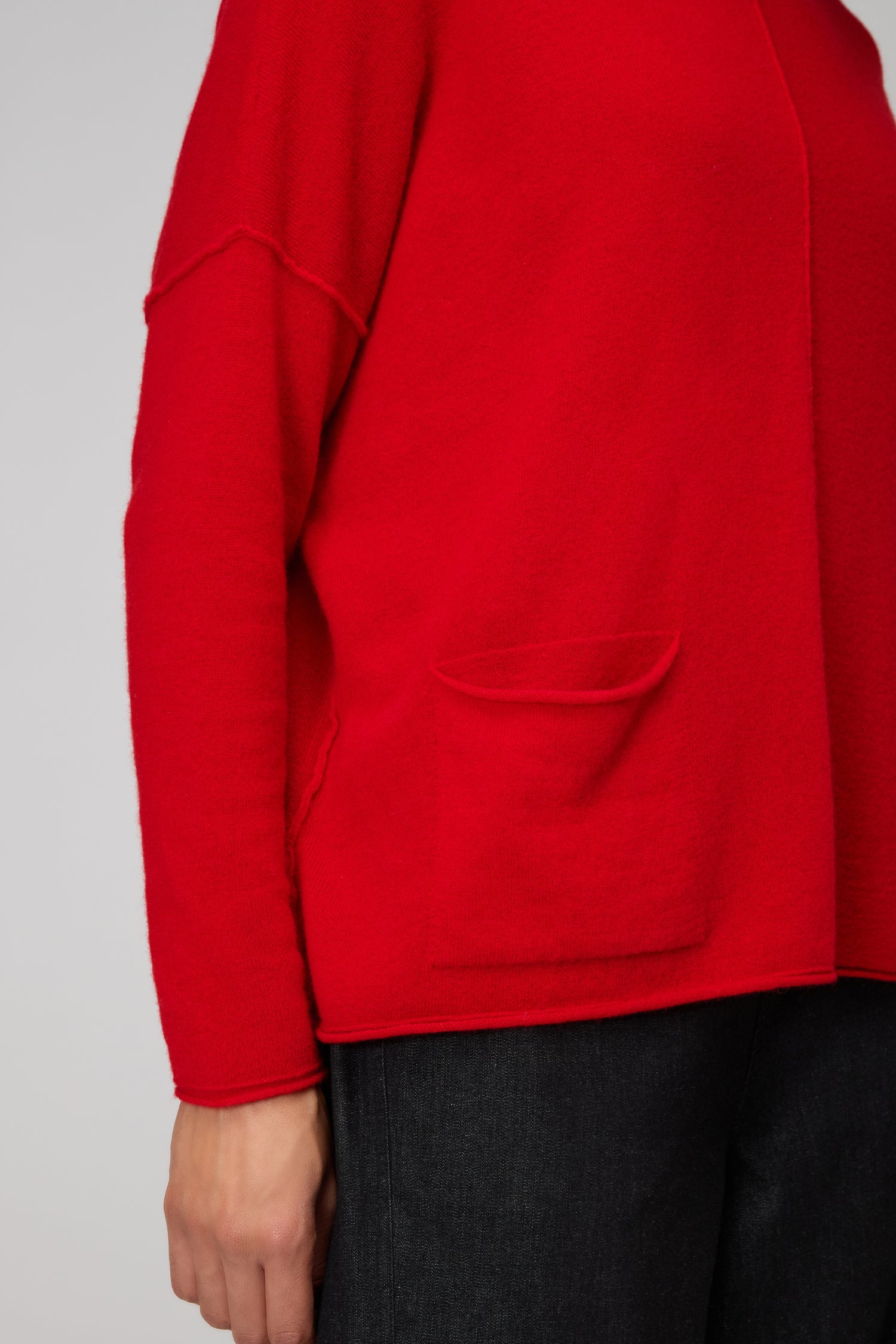 Someone wearing the Merino Pocket Jumper in bright red, featuring extra-fine merino wool and a pocket, paired with dark pants; shown from shoulder to hip against a plain background.