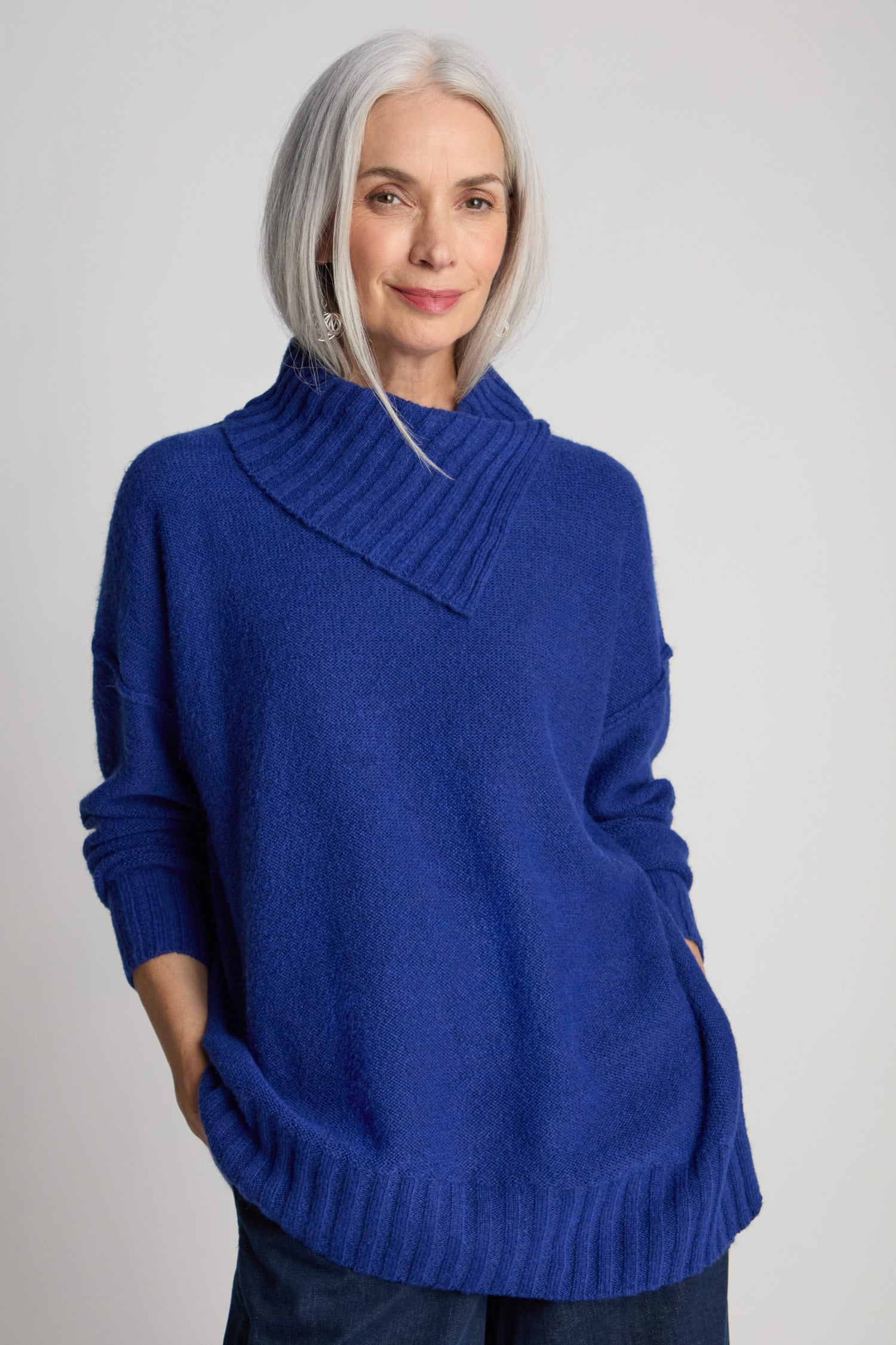 A woman with gray hair is wearing a contemporary-style Wool Viscose Mix Asymmetric Jumper in blue, which features a textured collar. She stands against a plain background with her hands in her pockets, smiling slightly.