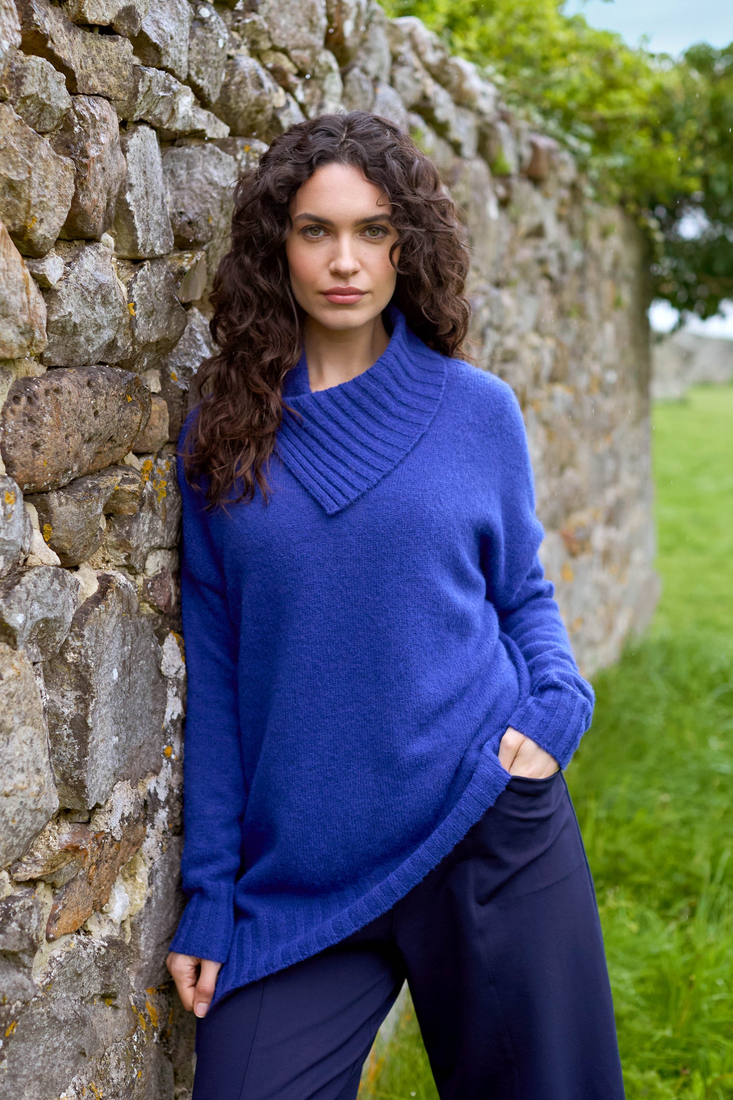 A person with long curly hair in a Wool Viscose Mix Asymmetric Jumper stands against a stone wall, with grass and trees in the background.