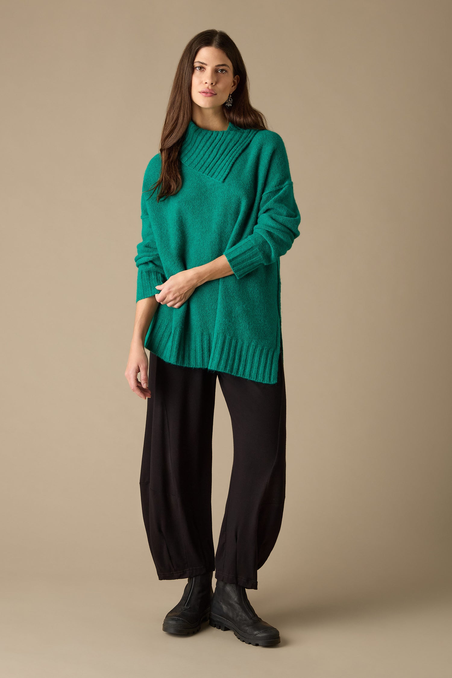 A person stands against a neutral background, showcasing a modern vibe in an Asymmetric Knit Jumper paired with black pants and boots, holding a pen.