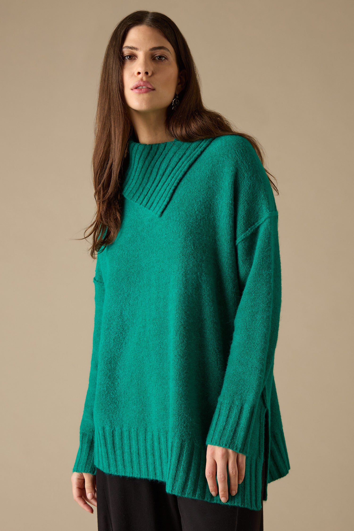 A person with long brown hair is wearing the Asymmetric Knit Jumper in teal with contemporary styling, paired with black pants, while standing against a beige background.