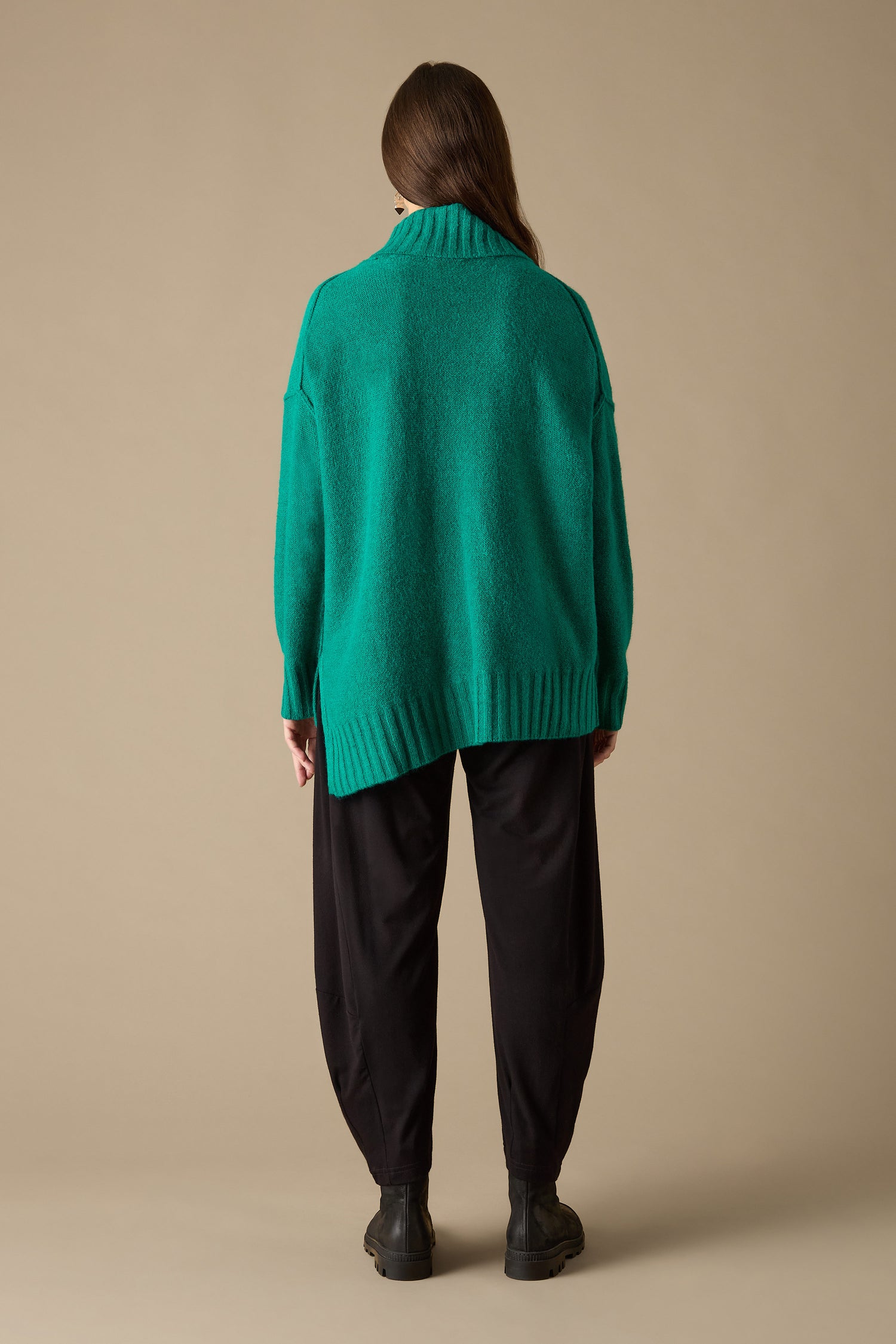 An individual is seen from behind wearing the Asymmetric Knit Jumper, exuding a contemporary style paired with black pants and boots against a simple backdrop.