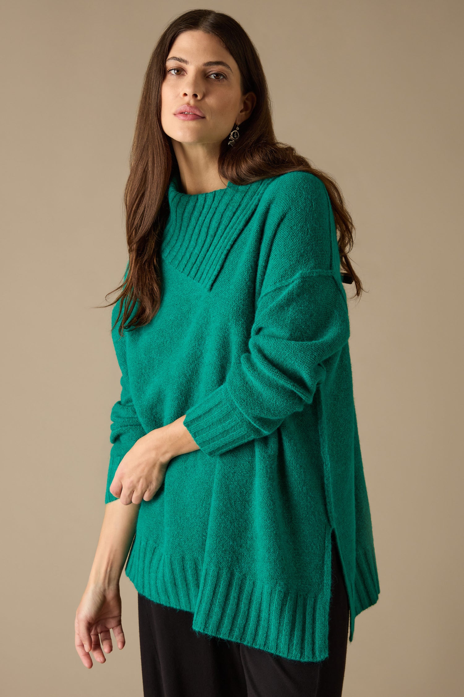 A person wearing the Asymmetric Knit Jumper in green, paired with black pants, stands against a plain background, showcasing a contemporary style with ribbed details and an oversized fit.