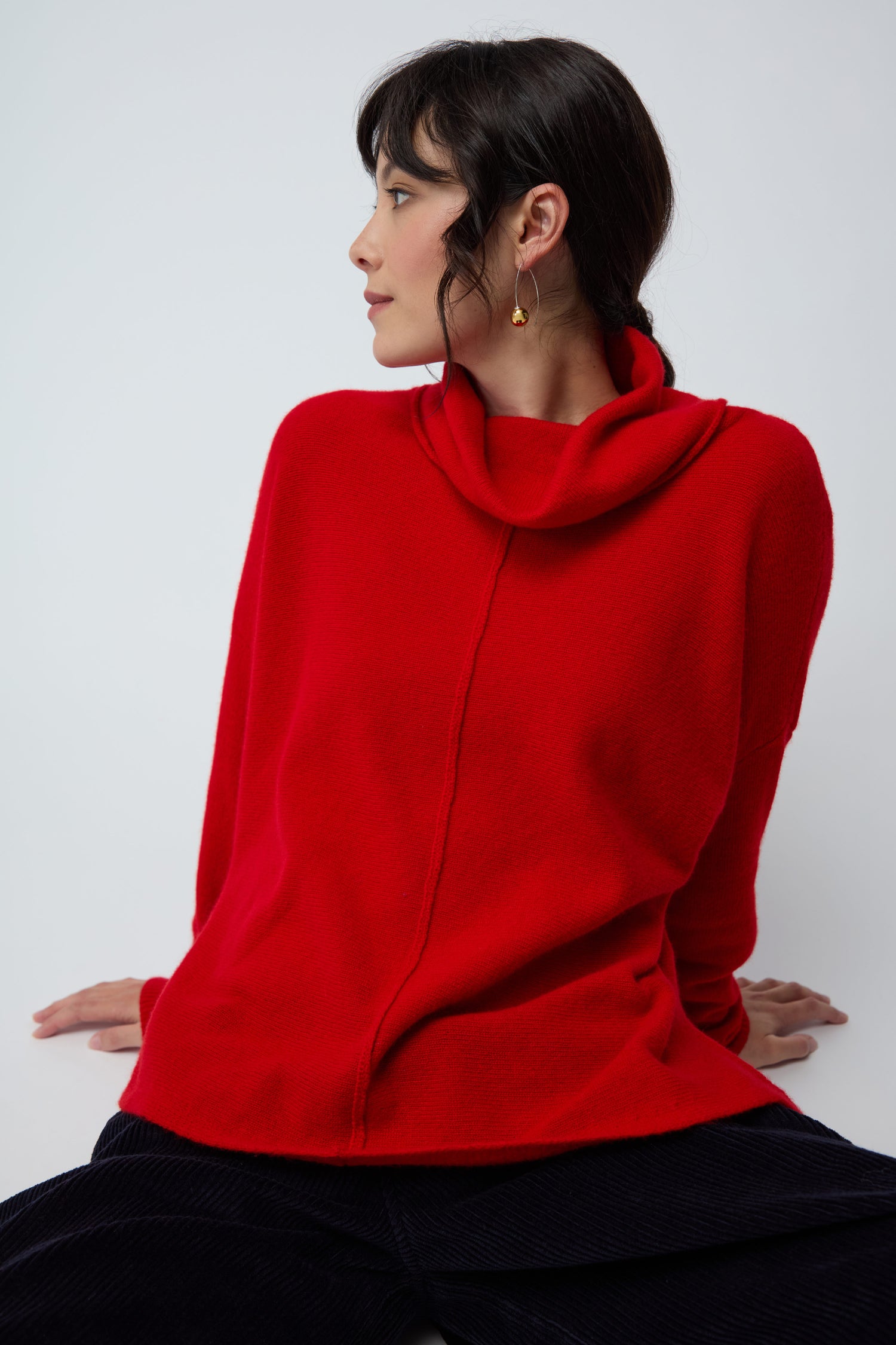 Woman seated against a plain background, wearing the Merino Wool Cowl Neck Jumper in bright red, dark pants, and gold hoop earrings, looking to her left.