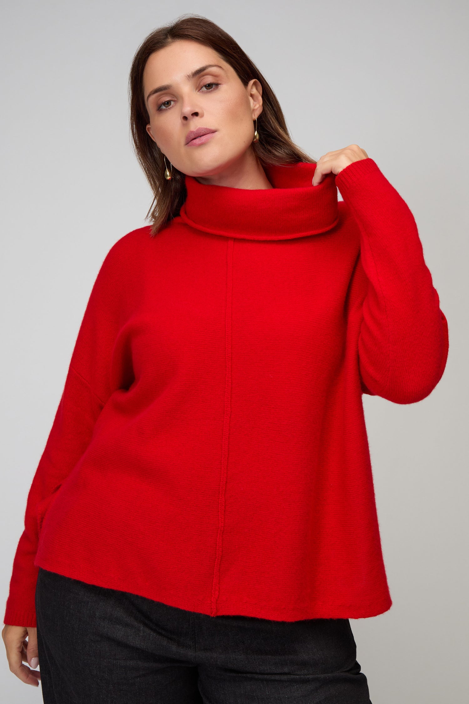 A person models the Merino Wool Cowl Neck Jumper in bright red with dark pants, posing against a plain light grey background.