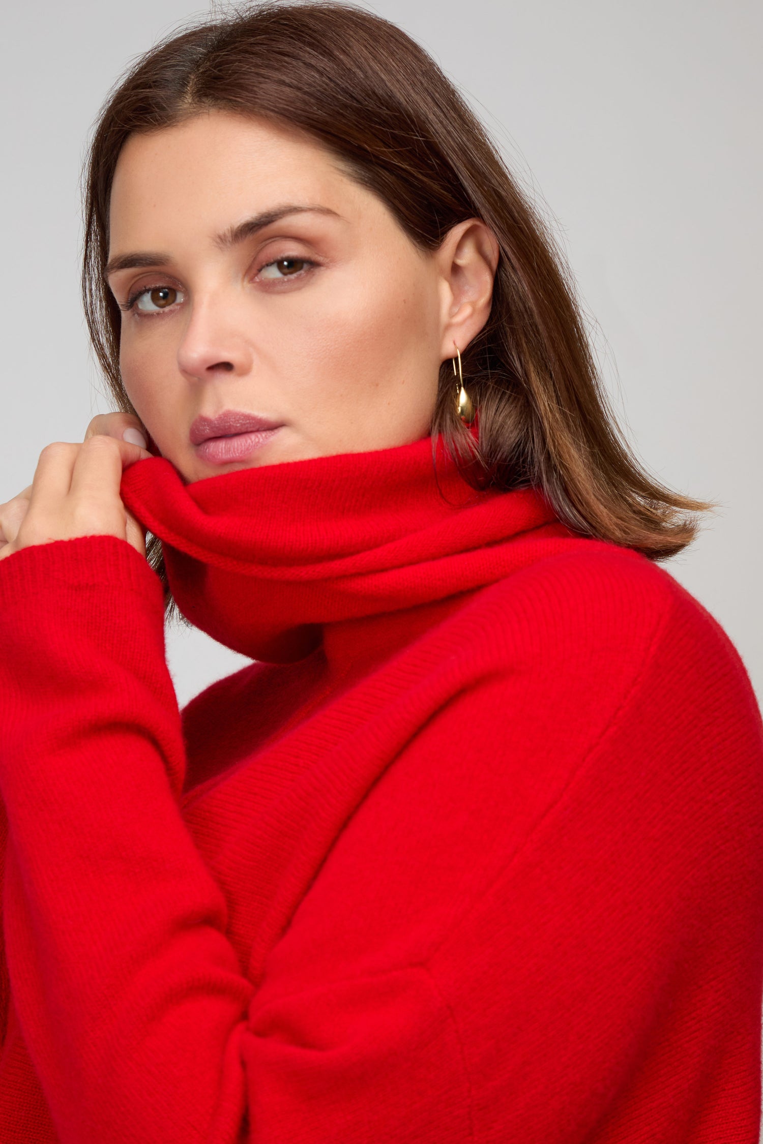 A woman with straight brown hair wears a bright red Merino Wool Cowl Neck Jumper and gold hoop earrings, looking directly at the camera.