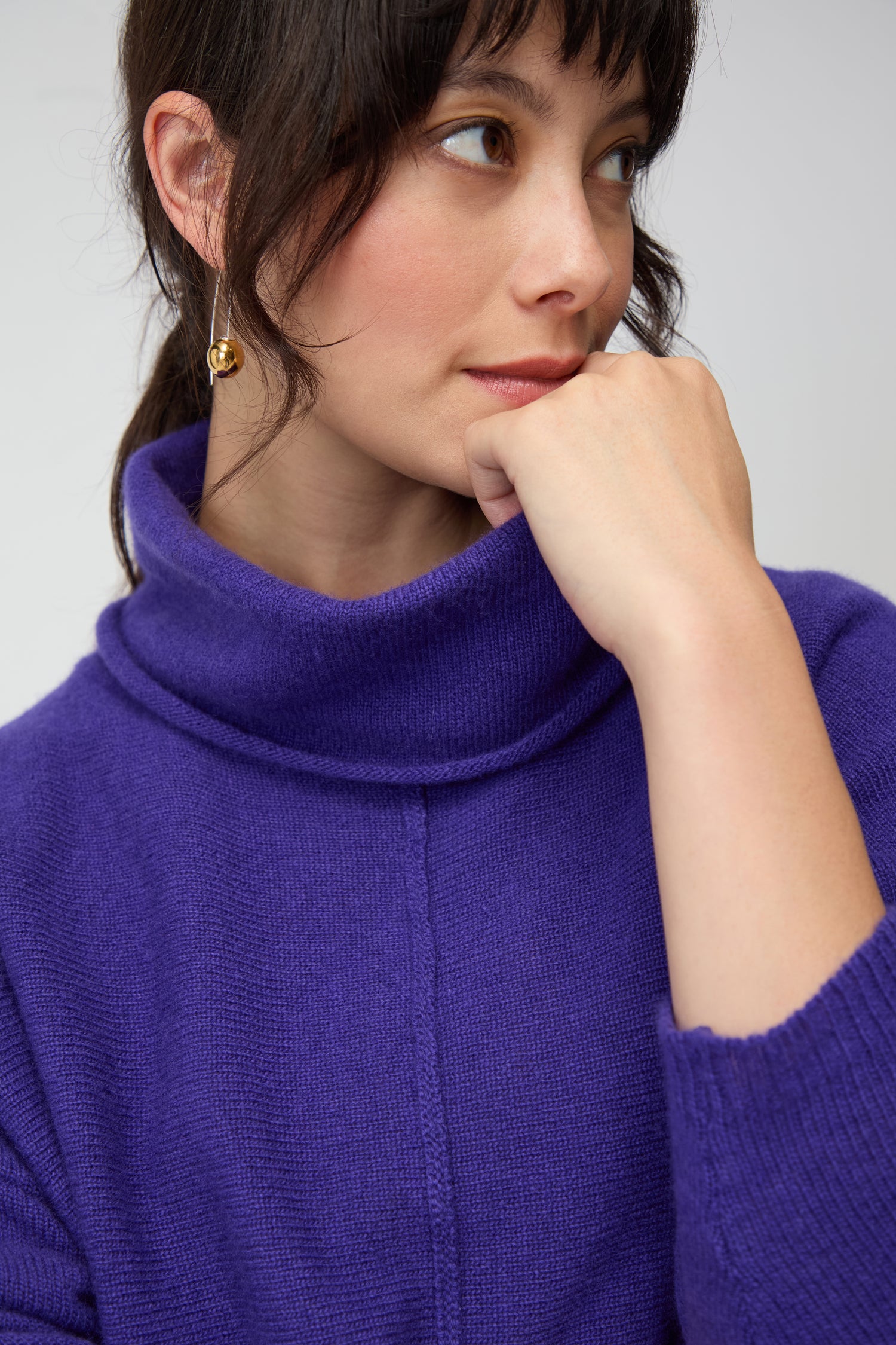 A woman in a Merino Wool Cowl Neck Jumper rests her chin on her hand and gazes to the side.