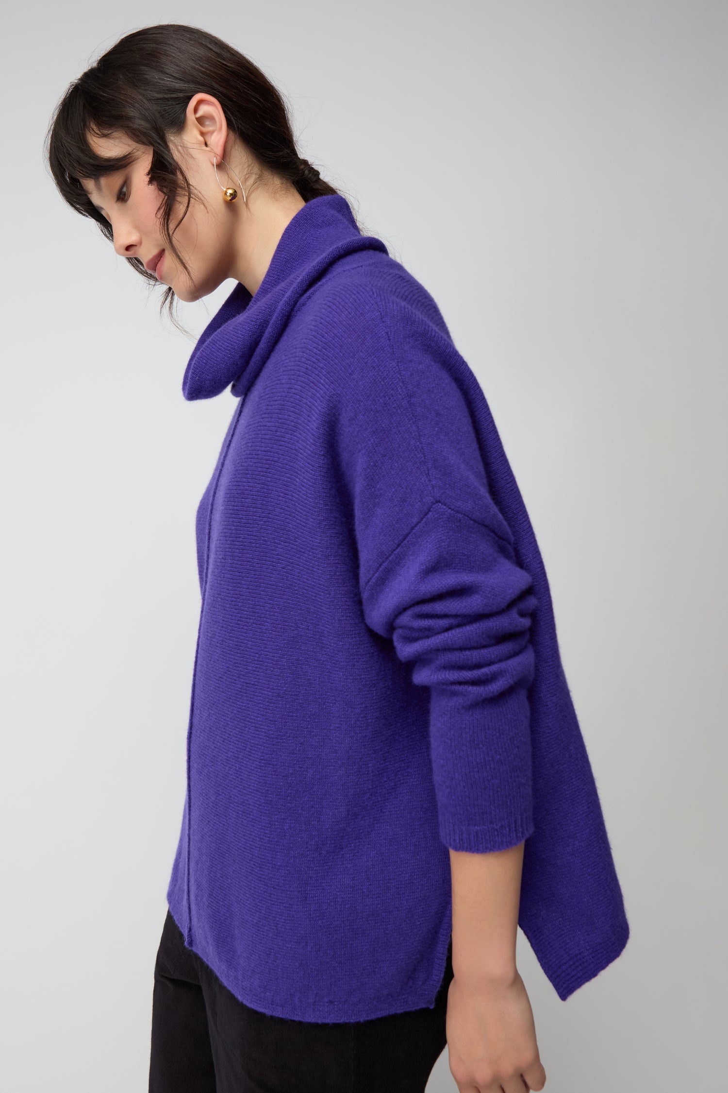 A person with dark hair wears a purple Merino Wool Cowl Neck Jumper and black pants, standing against a plain light gray background.