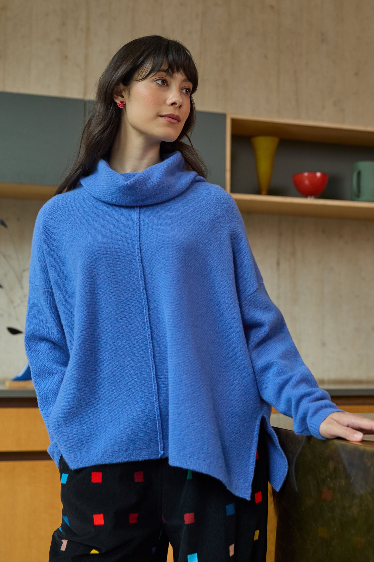 A woman stands indoors by shelves and kitchenware, wearing the Merino Cowl Neck Jumper in loose blue wool and black pants with colorful square patterns.