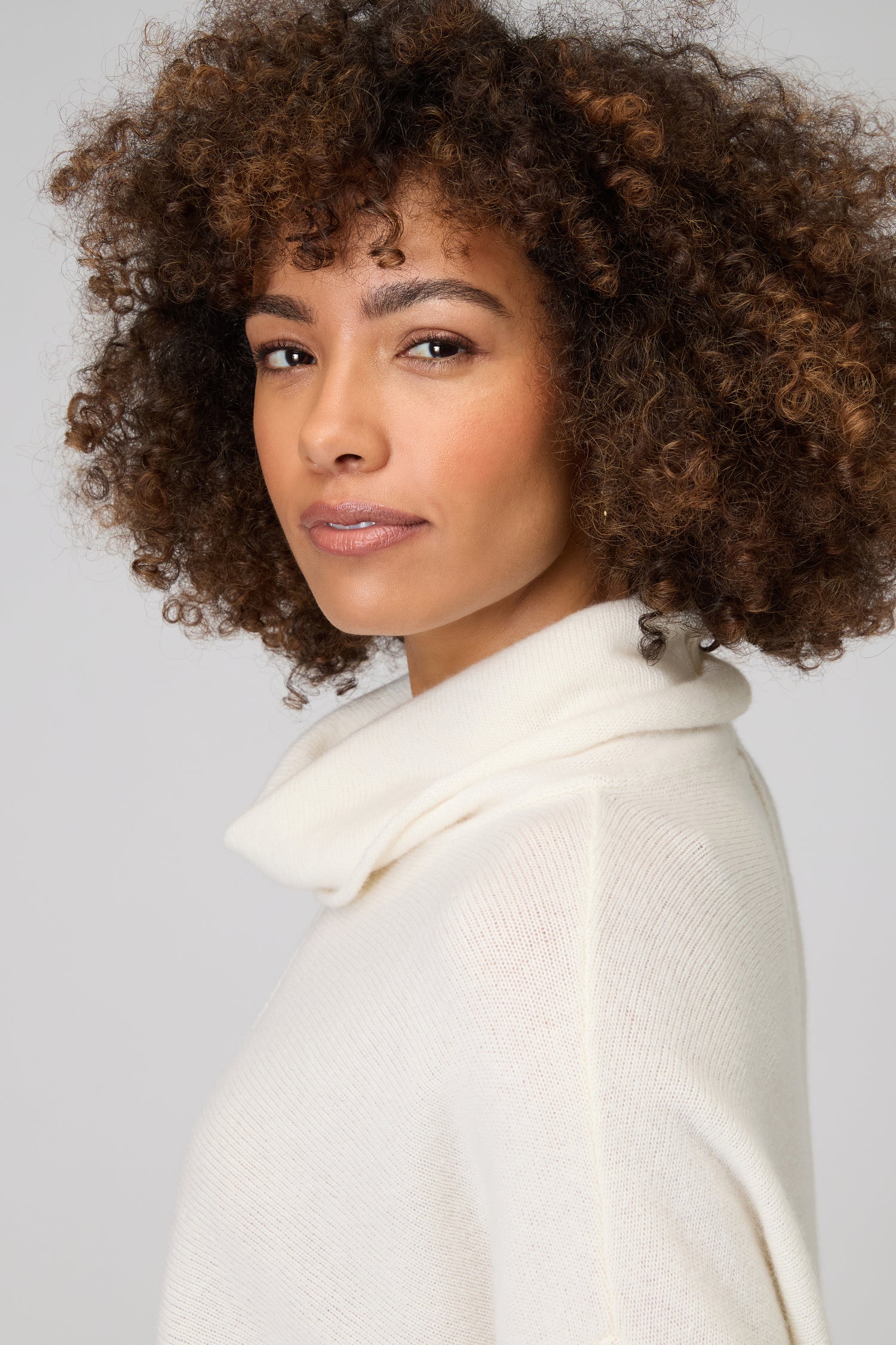 A woman with curly hair wears the Merino Cowl Neck Jumper in off-white, looking at the camera with a neutral expression against a plain background.