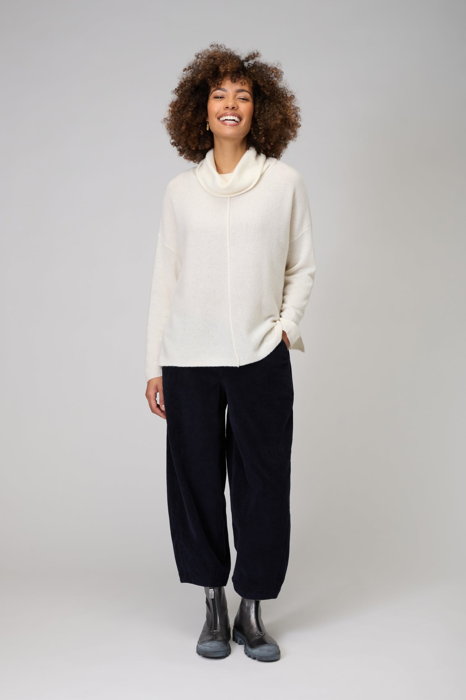 A woman stands against a plain background, smiling with one hand in her pocket, wearing the Merino Cowl Neck Jumper in cream alongside loose black pants and gray boots—an elegant showcase of women's luxury knitwear.