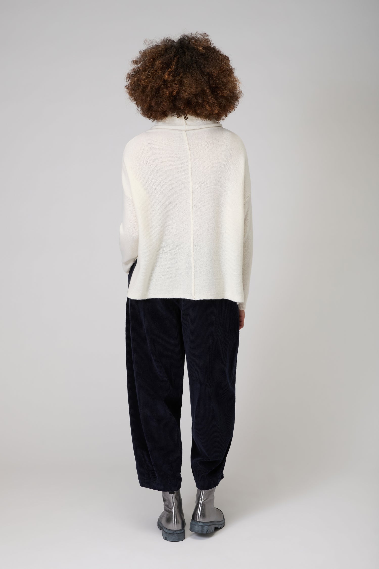 A person stands facing away against a plain light background, wearing the Merino Cowl Neck Jumper in cream, paired with loose dark pants and gray ankle boots.