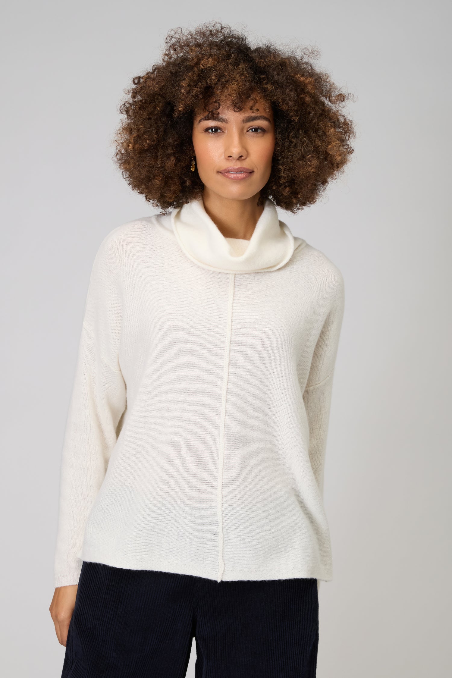 A person with curly hair wears the Merino Cowl Neck Jumper, a women’s luxury knitwear piece in cream, paired with dark pants and standing against a plain light gray background.