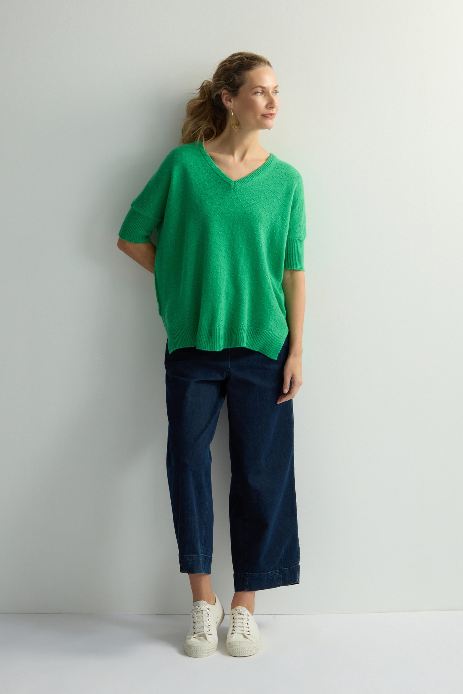 A person stands against a white wall, wearing a Merino V Neck Knot Stitch Jumper in green, dark pants, and white shoes. They gaze thoughtfully to their right.