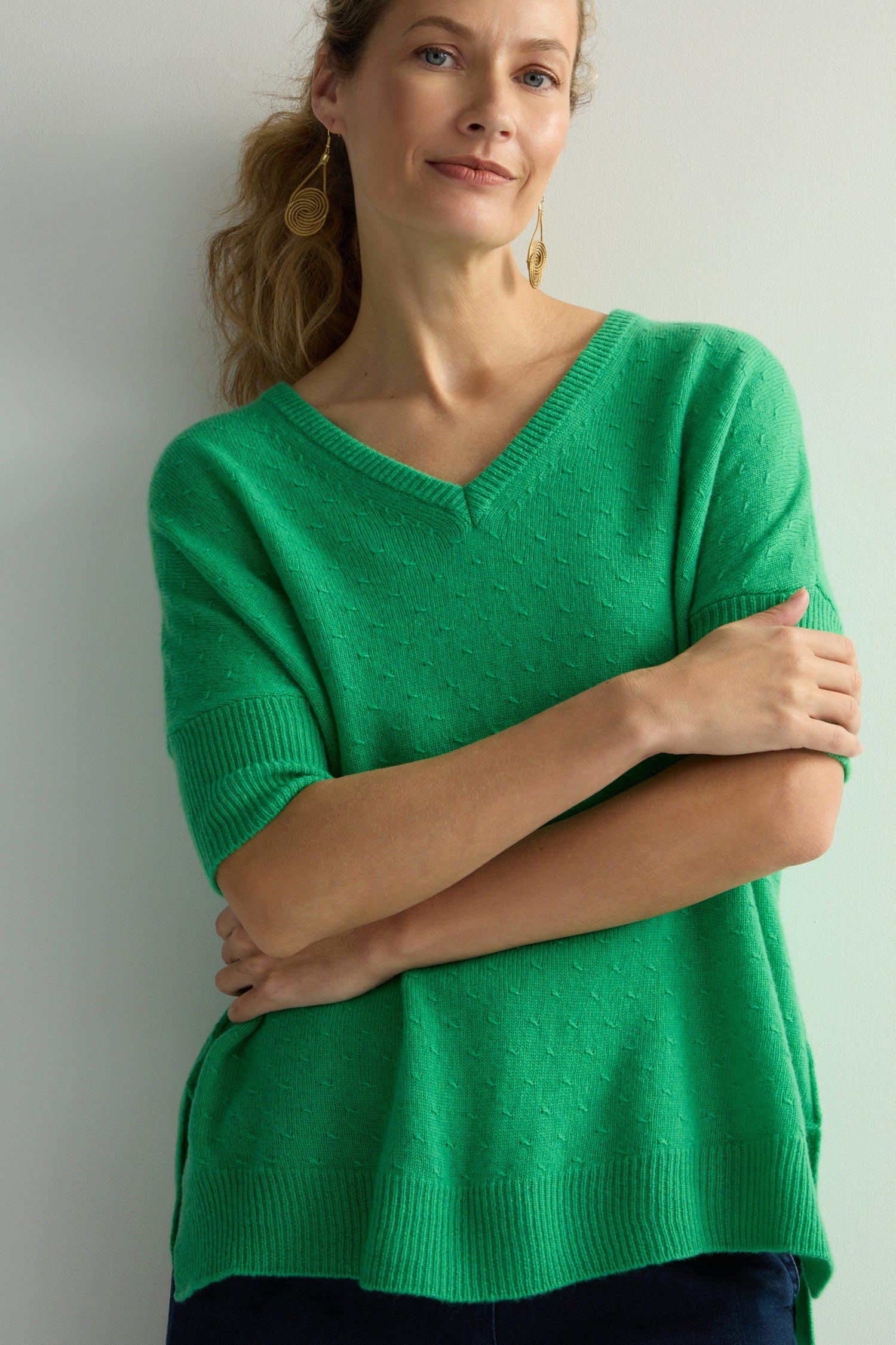 A person stands against a plain background with arms crossed, wearing a green Merino V Neck Knot Stitch Jumper.