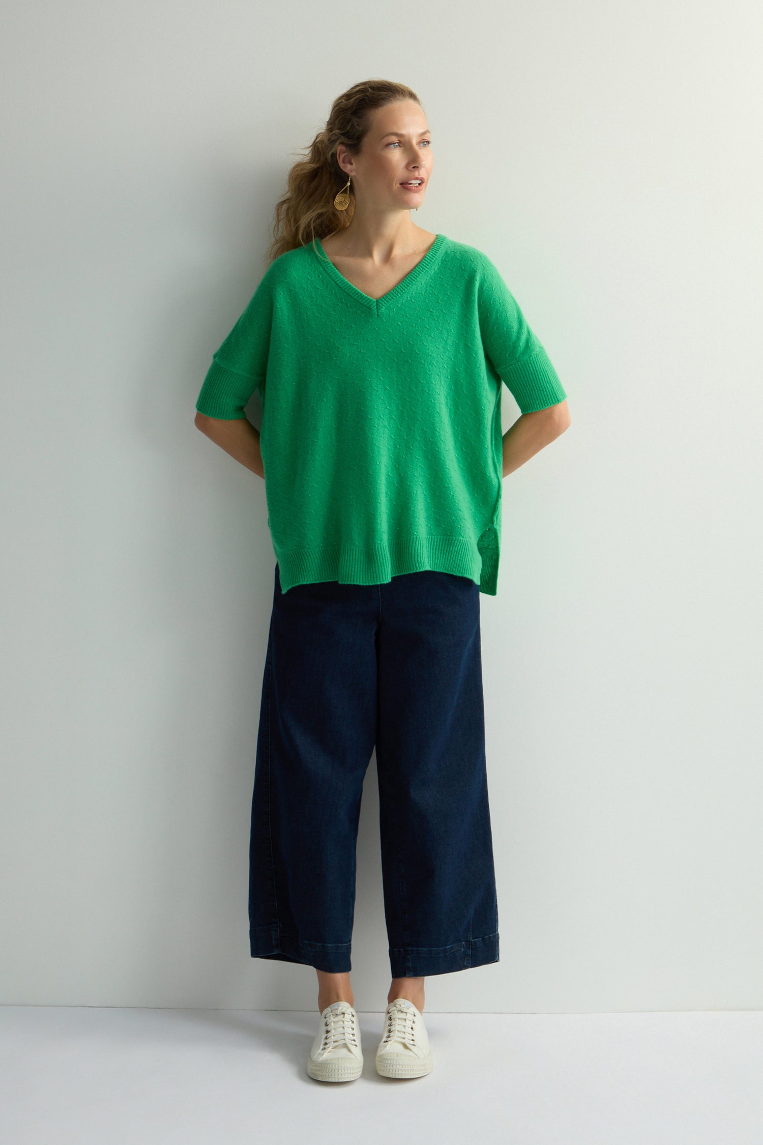 A person stands against a light background wearing a Merino V Neck Knot Stitch Jumper in green, dark wide-leg pants, and white sneakers. They have wavy hair and are looking to the side.