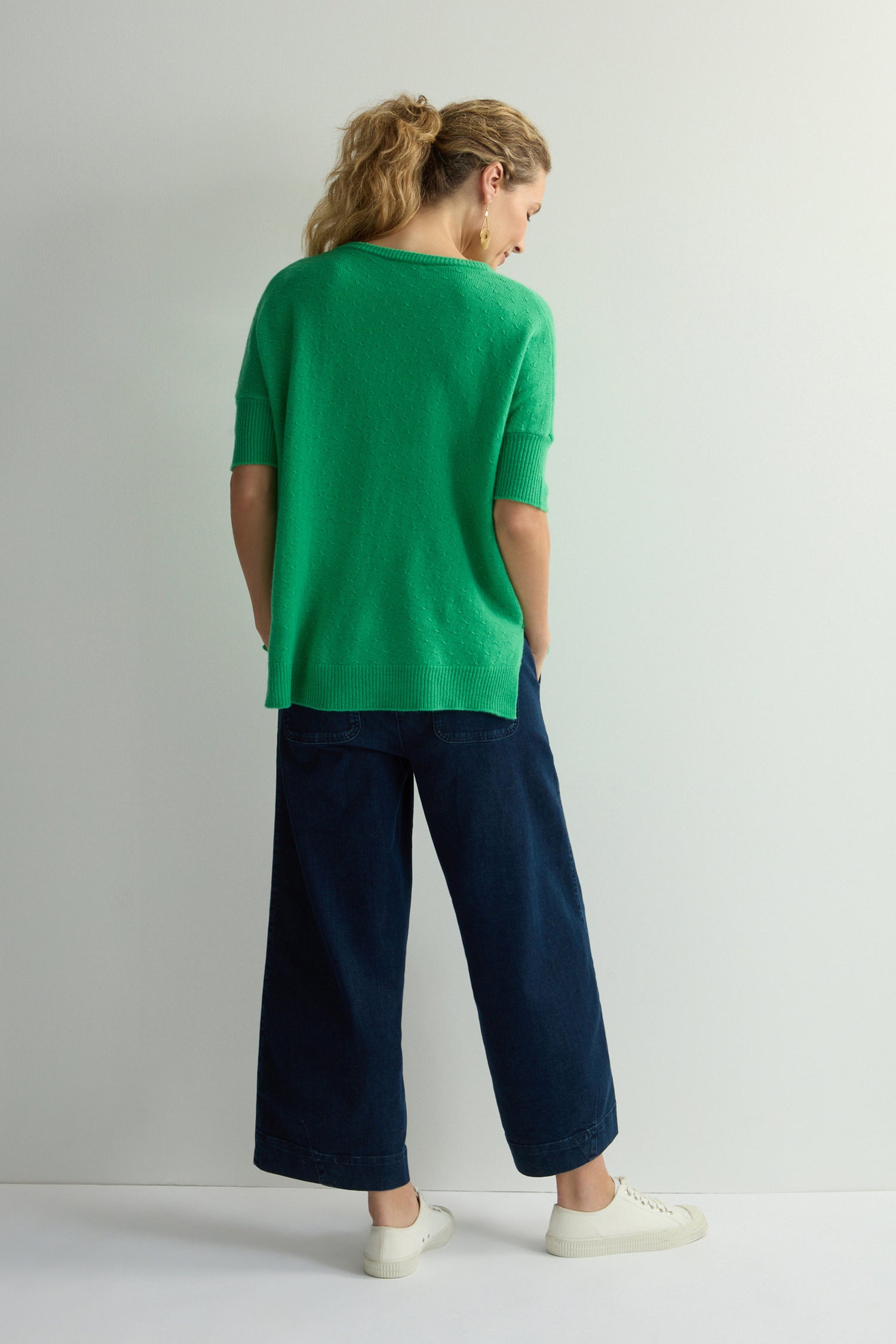 A person wearing a Merino V Neck Knot Stitch Jumper and navy wide-leg pants stands against a light background.
