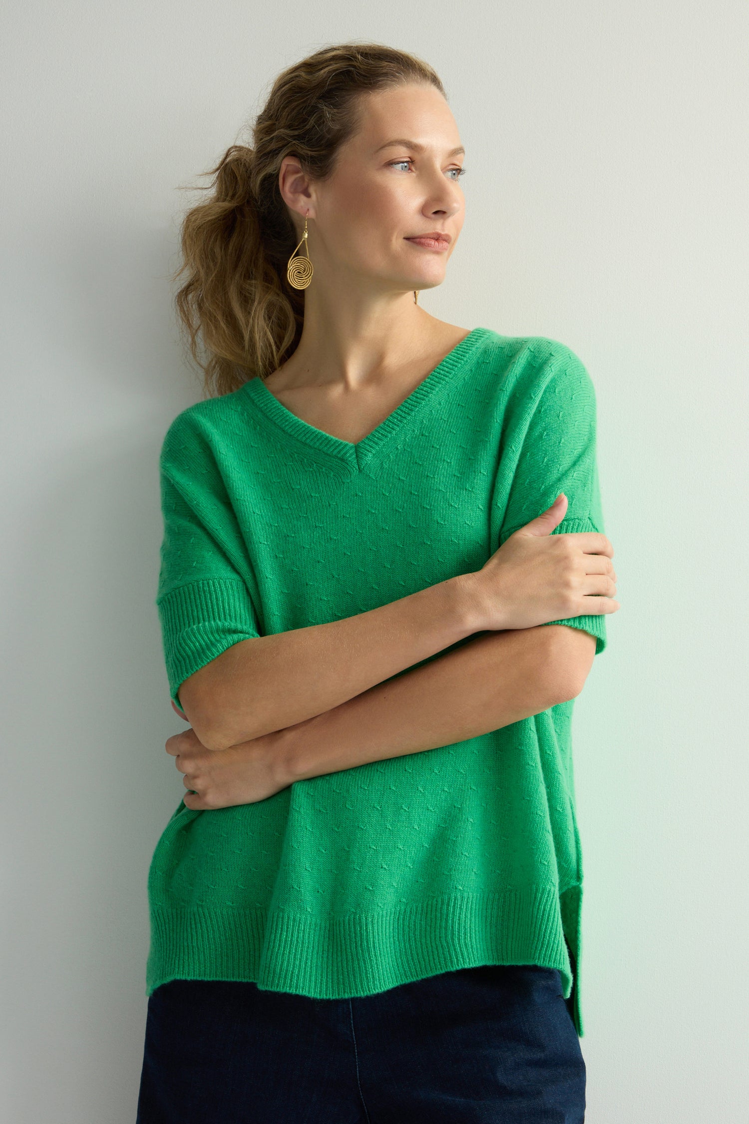 A person in a Merino V Neck Knot Stitch Jumper stands against a light wall, arms crossed, looking to the side while wearing gold earrings.