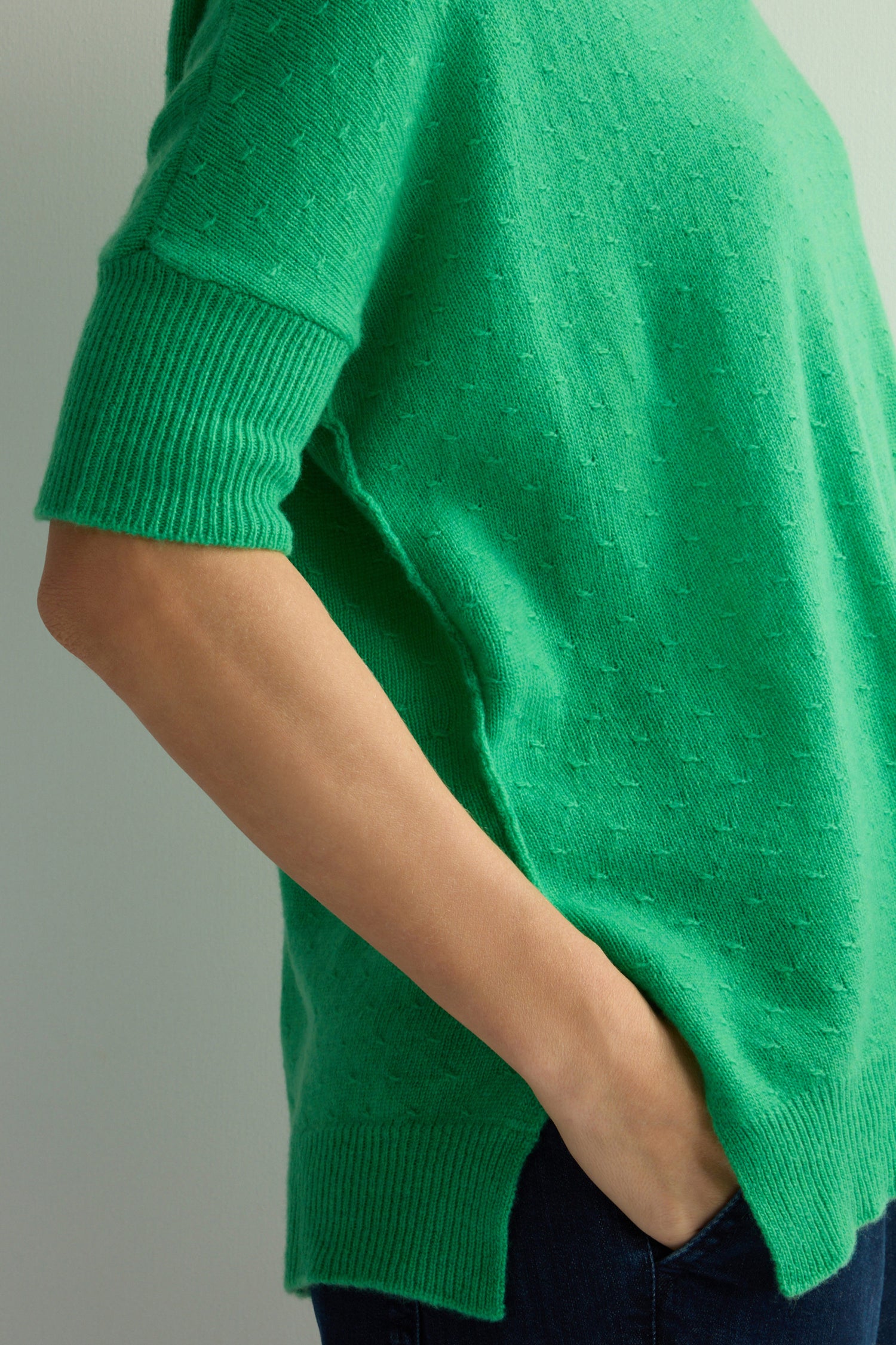 Individual wearing a green Merino V Neck Knot Stitch Jumper with hands in jeans pockets against a neutral background.