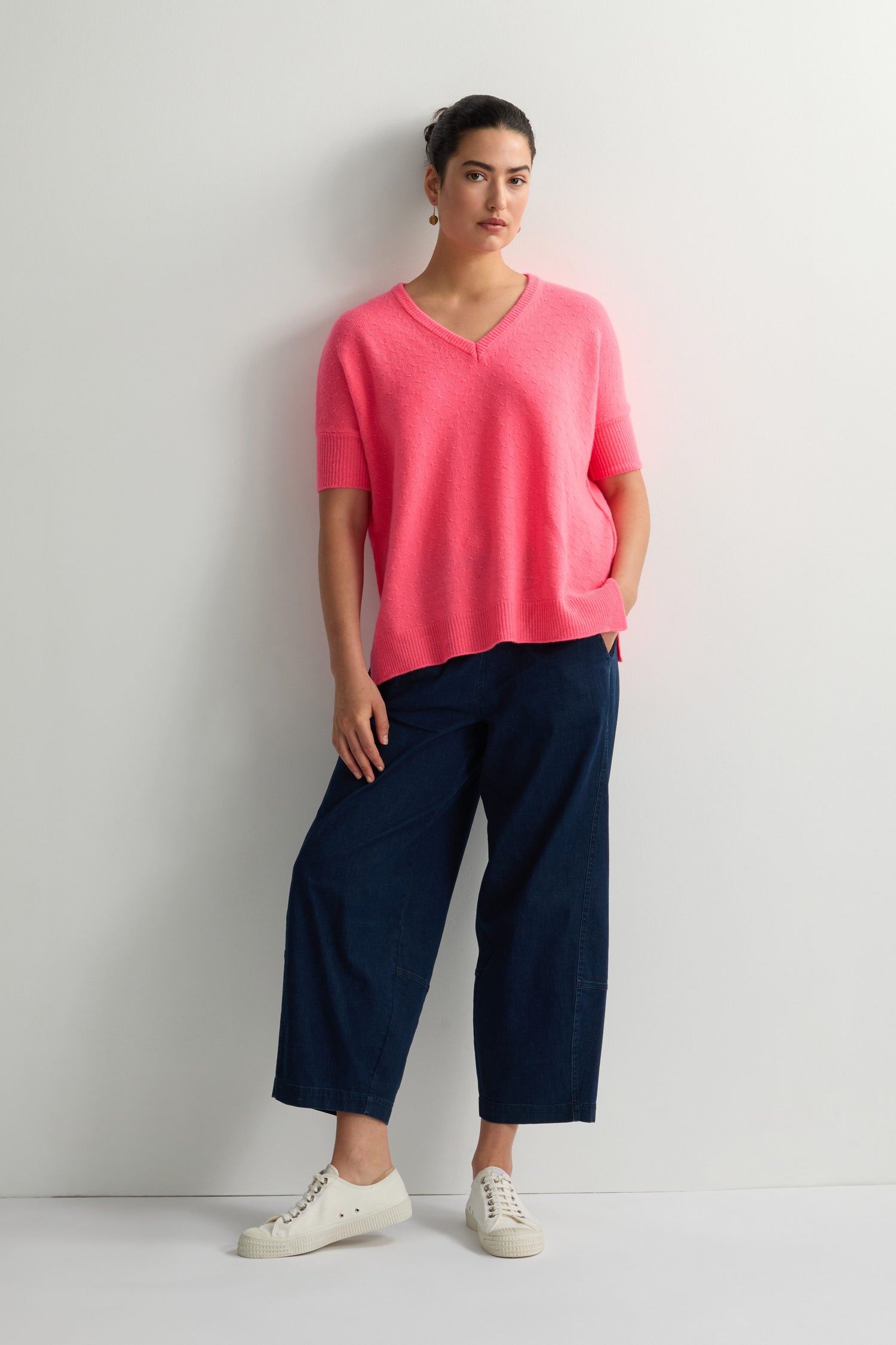 A person stylishly wears a bright pink Merino V Neck Knit Stitch Jumper, paired with dark wide-leg pants and white sneakers, against a plain white wall.