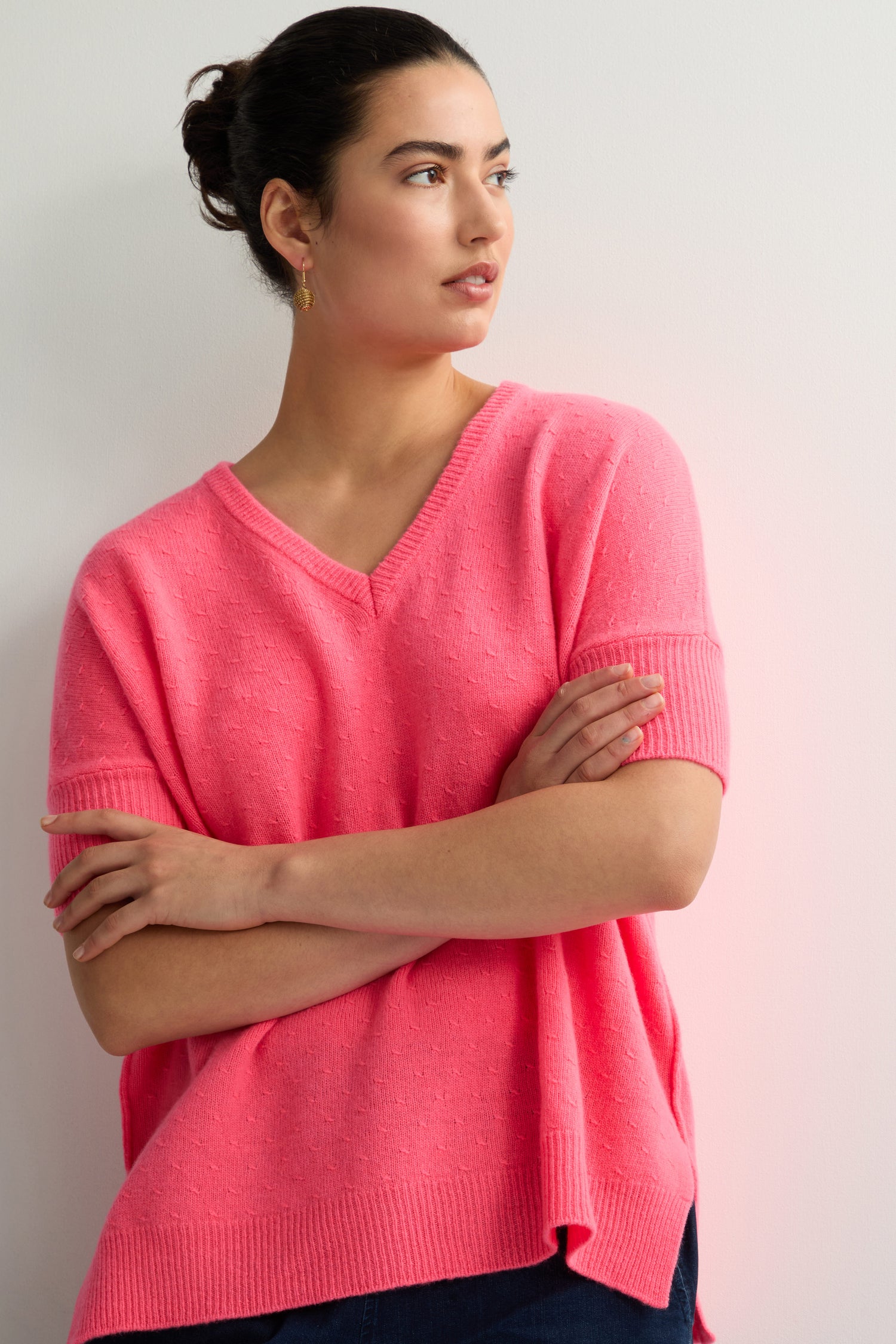 A person with dark hair in a bun, wearing a pink "Merino V Neck Knot Stitch Jumper" and hoop earrings, stands against a light-colored wall. They're looking to the side with arms crossed, exuding confidence.