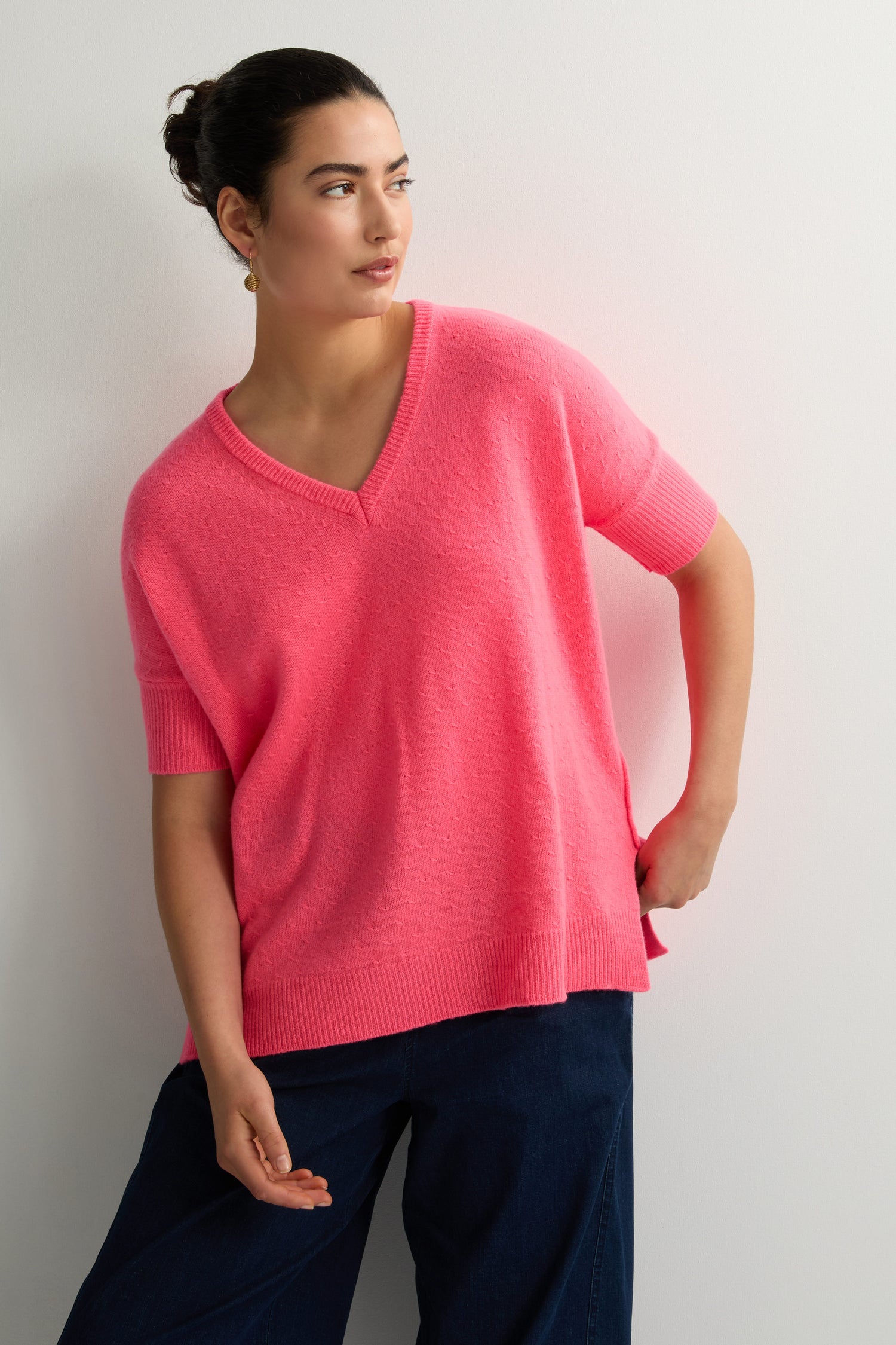 A person wearing a Merino V Neck Knot Stitch Jumper in pink and blue pants stands against a plain white wall, looking to the side.