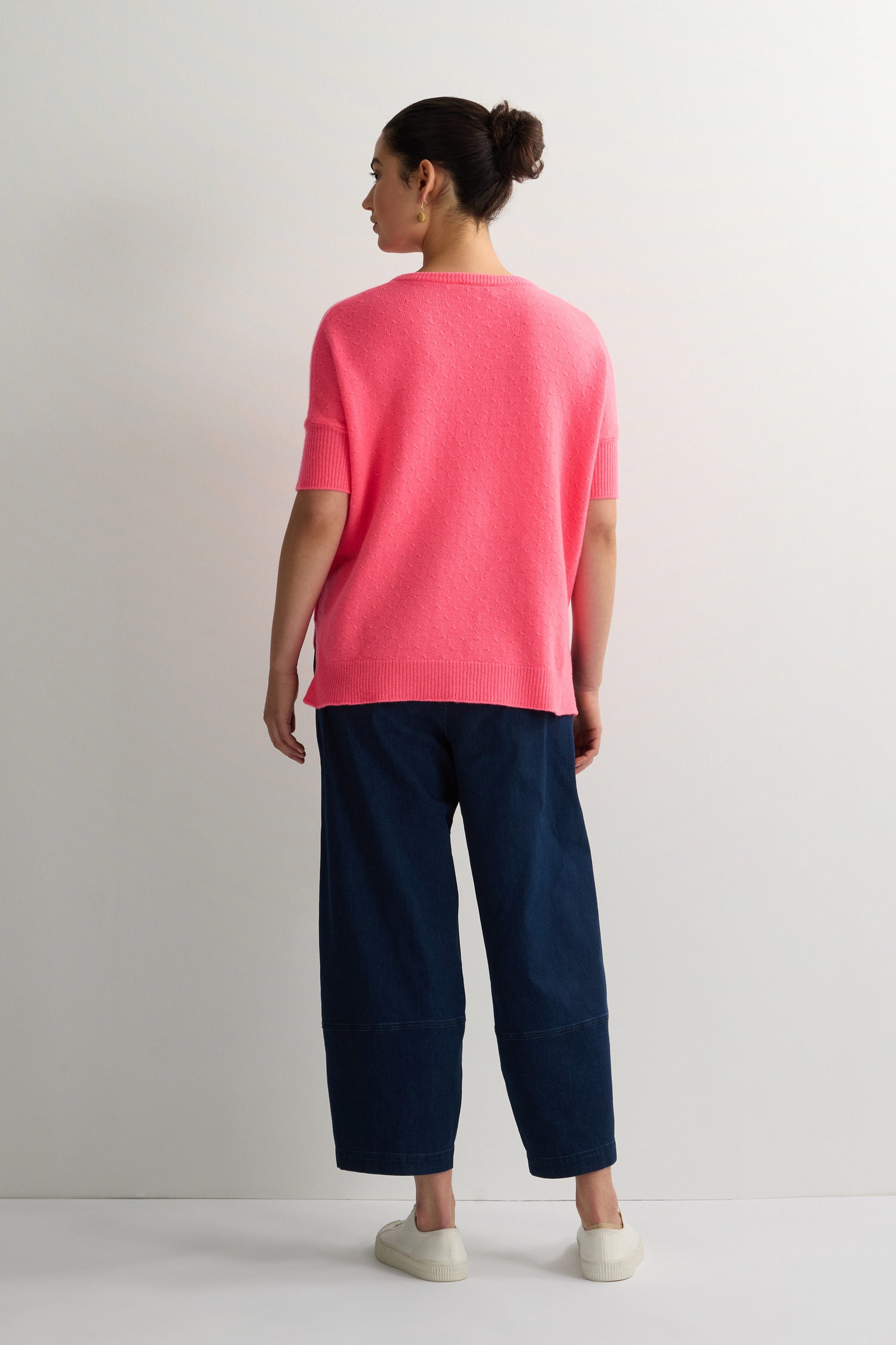 A person faces away in a plain white room, wearing a bright pink Merino V Neck Knot Stitch Jumper, dark wide-leg pants, and white slip-on shoes.