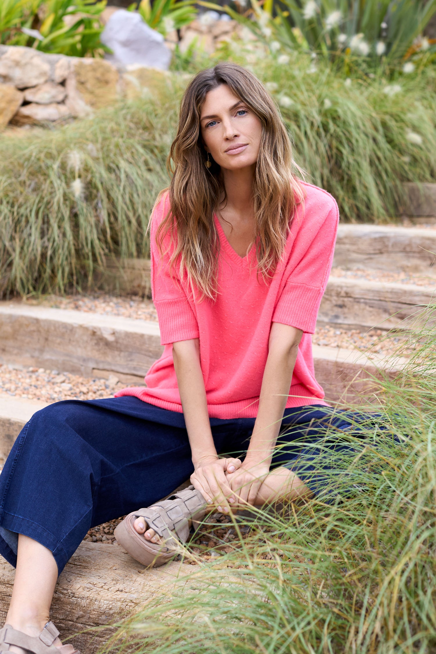 A woman with long hair sits on outdoor wooden steps, wearing a pink Merino V Neck Knot Stitch Jumper and dark wide-legged pants.