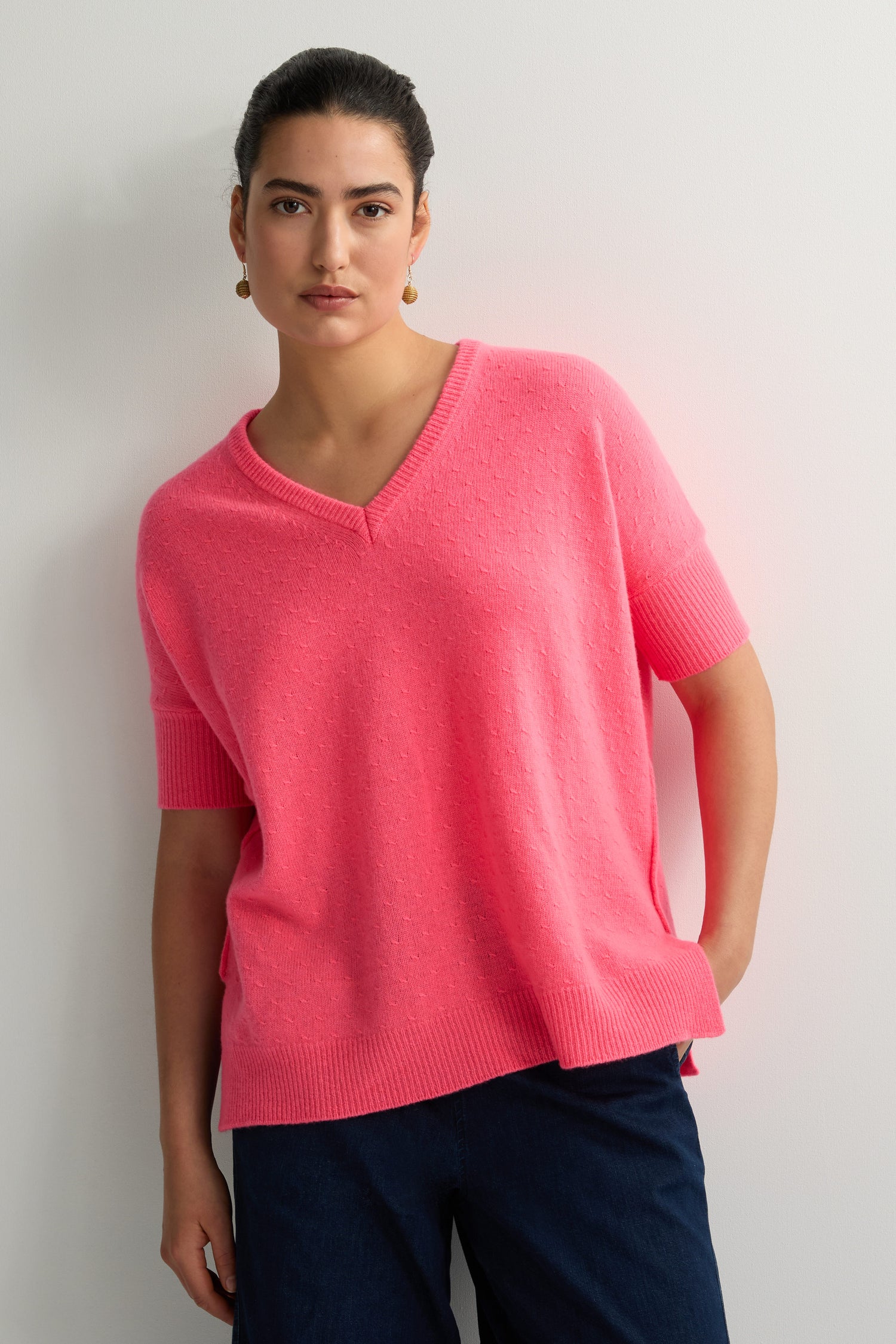 Person wearing a Merino V Neck Knot Stitch Jumper in pink and dark pants, standing against a light-colored wall.