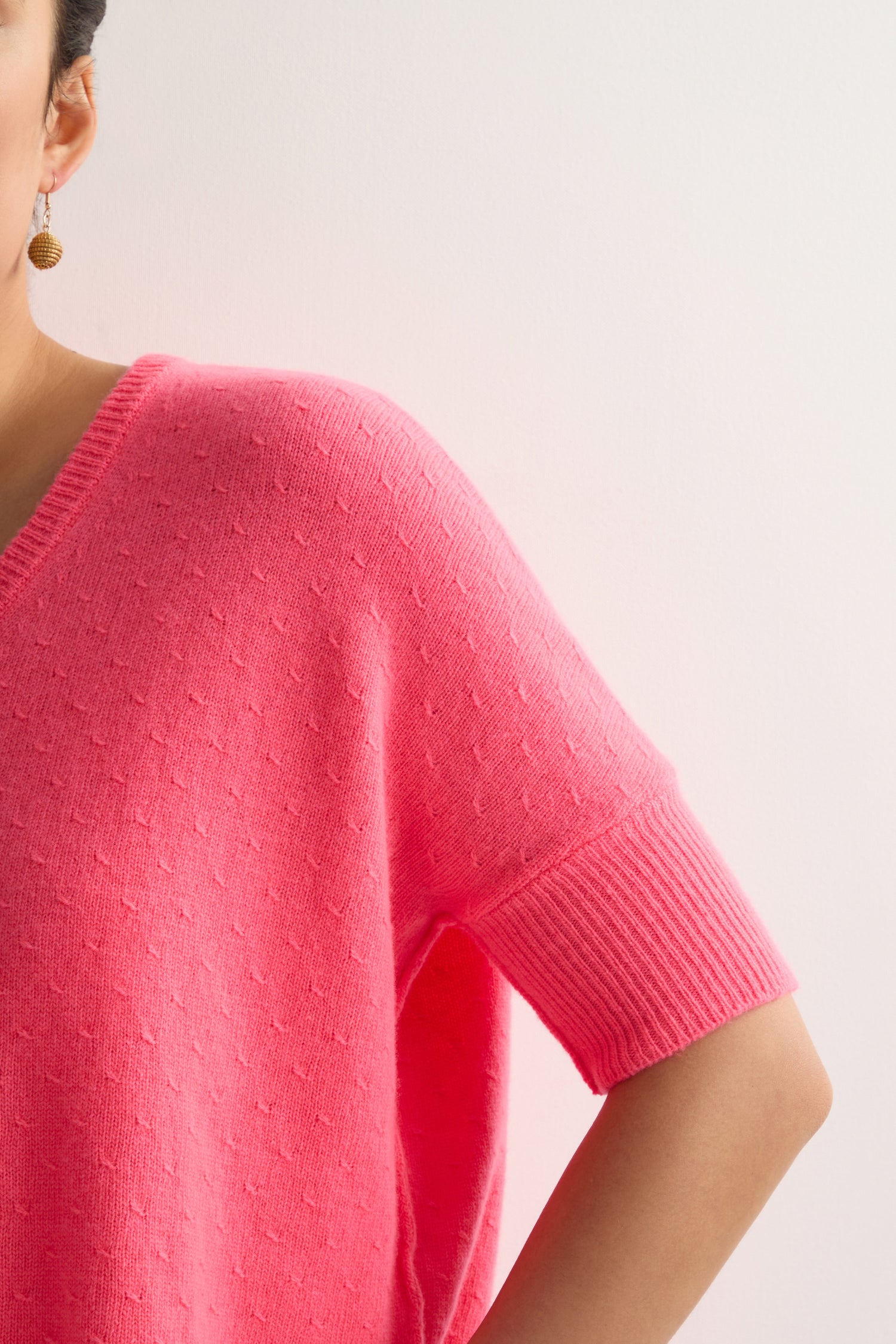 A person sports a Merino V Neck Knot Stitch Jumper in bright pink, showcasing just the right upper body and bent elbow. A partially visible gold hoop earring adds elegance to this cozy extrafine wool ensemble.