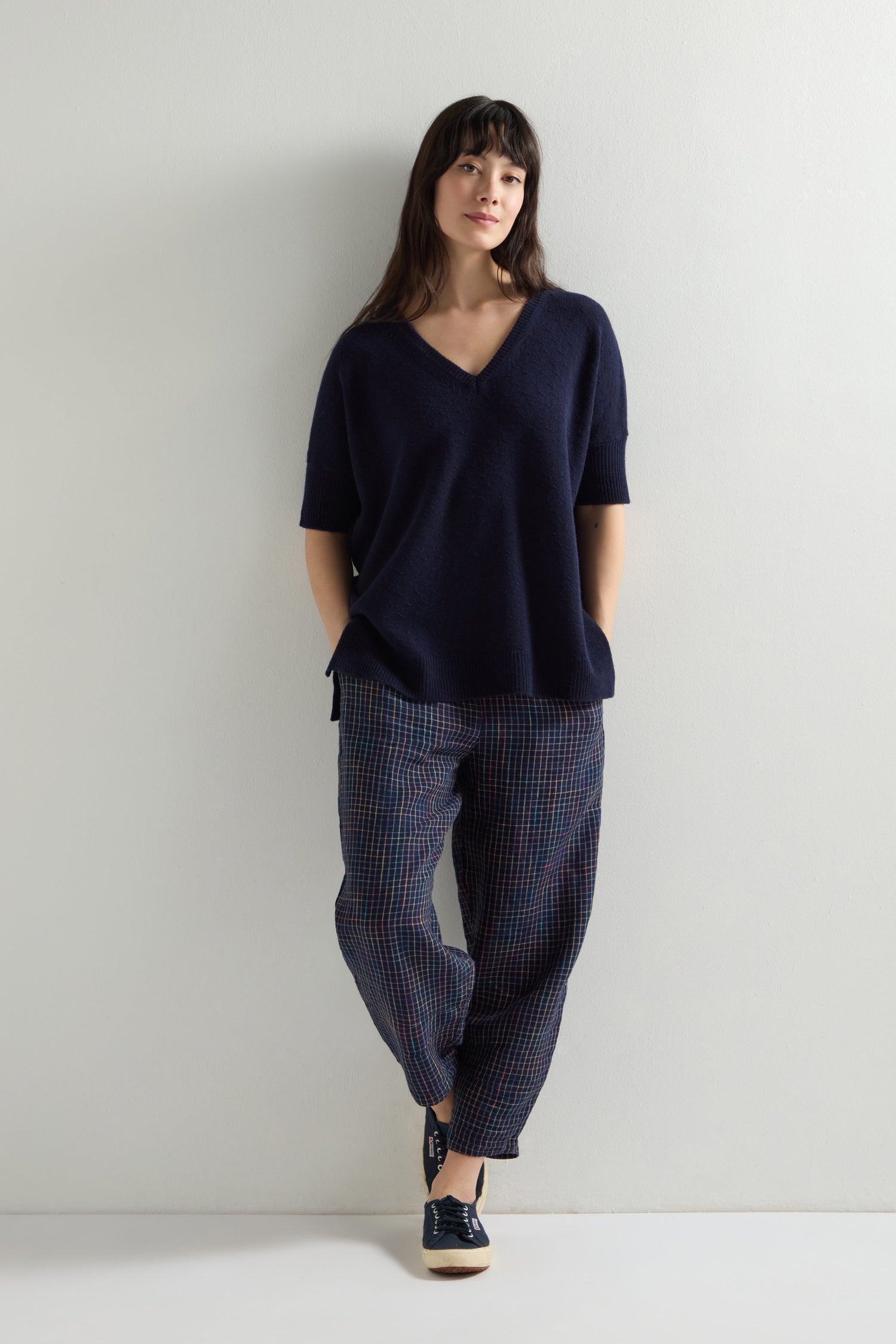 A person stands against a plain wall, wearing a Merino V Neck Knot Stitch Jumper. Their plaid pants add to the casual look, with hands in pockets, while the jumper's knot stitch detailing enhances their relaxed yet elegant style.